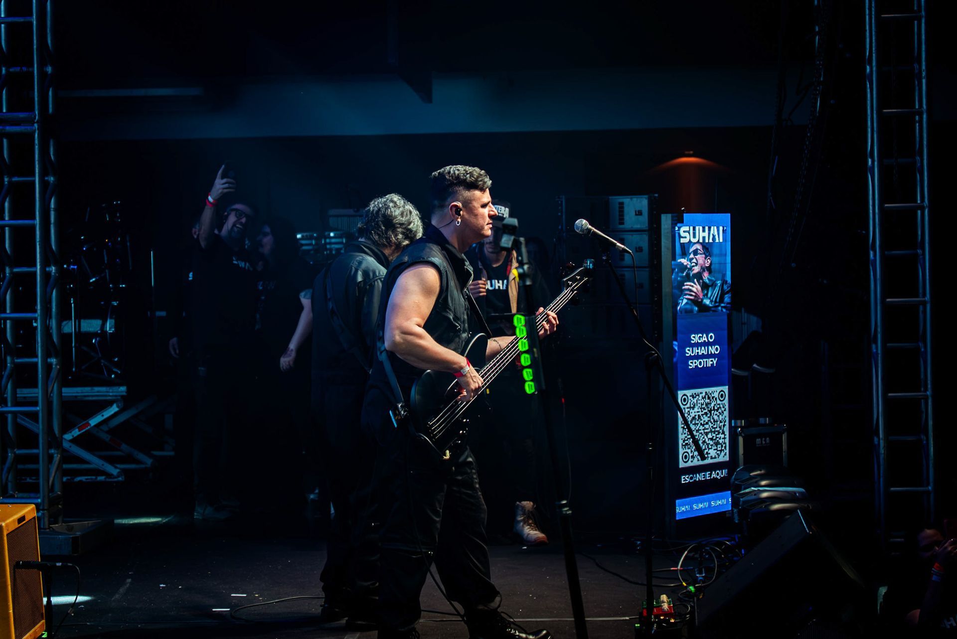 Um músico tocando um baixo elétrico em um palco escuro, ao lado de uma tela vertical que exibe um código QR.