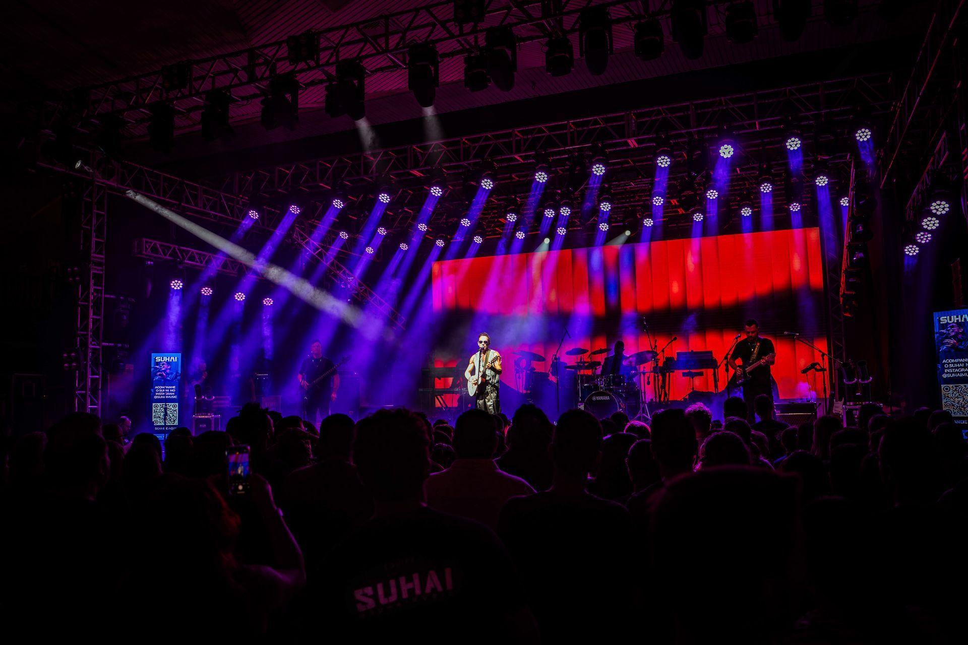 Um artista no palco tocando guitarra para uma plateia, iluminado por holofotes roxos vibrantes e um fundo vermelho.