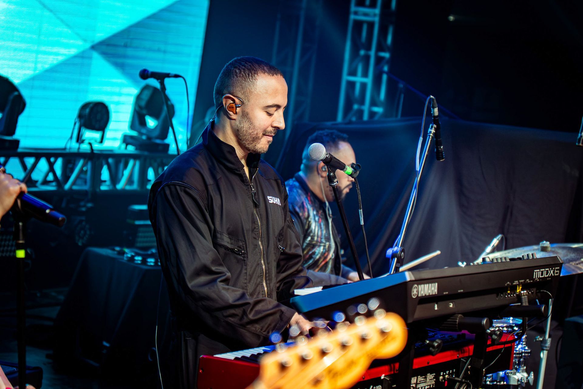 Dois músicos se apresentam no palco com teclados em um local com pouca luz e iluminação azul.