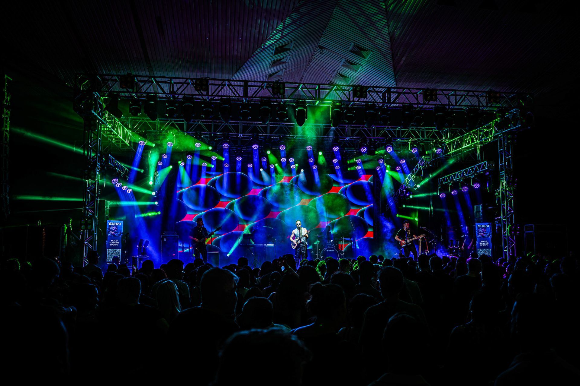 Um palco de concertos com músicos banhados por vibrantes holofotes azuis e verdes, vistos por trás de uma multidão escura.