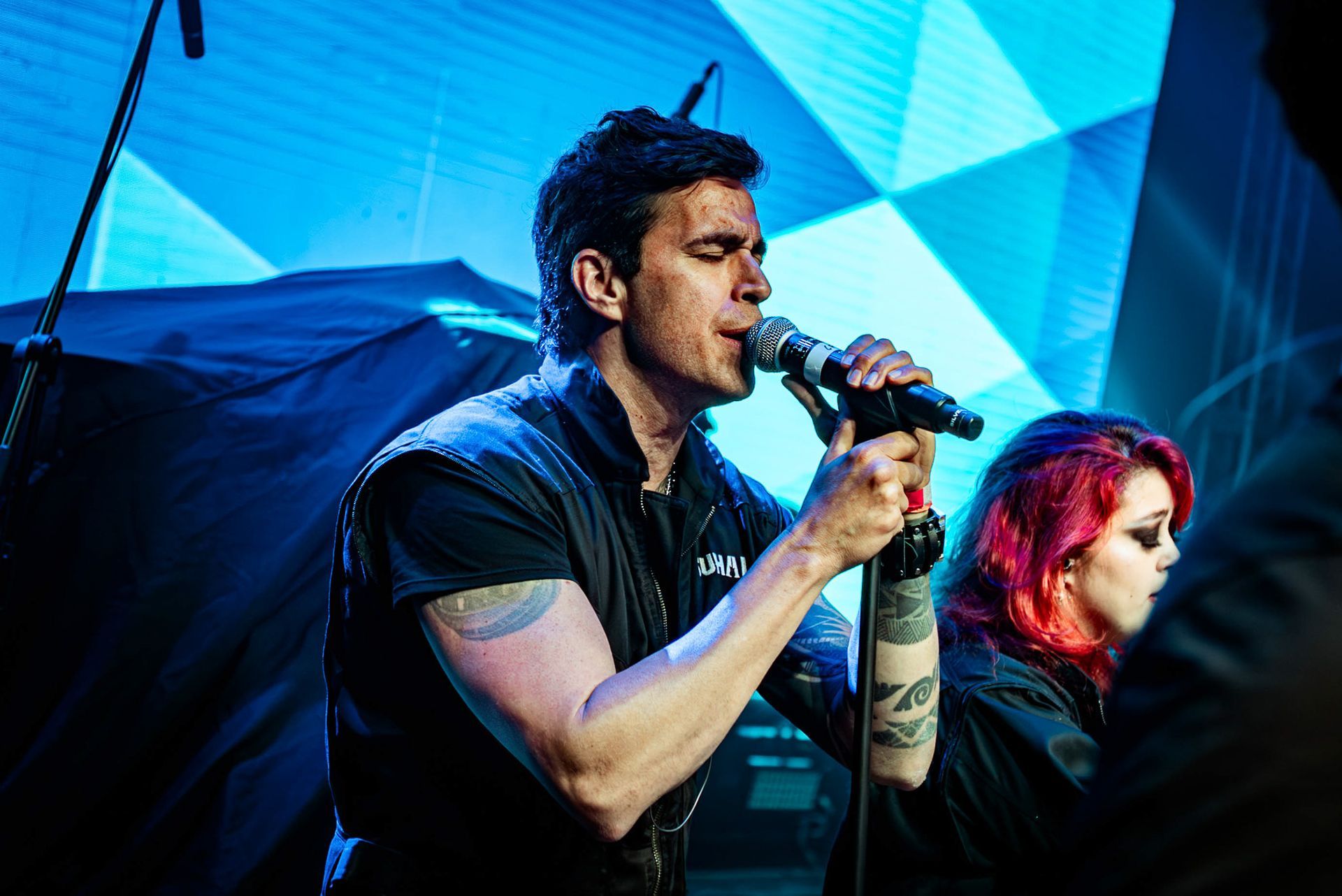 Um músico cantando ao microfone no palco, com uma pessoa de cabelos ruivos brilhantes parcialmente visível ao lado.