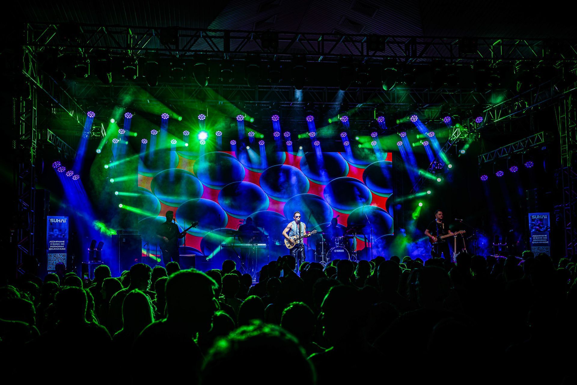 Um músico toca guitarra em um palco de concertos iluminado por luzes vibrantes em tons de azul e verde