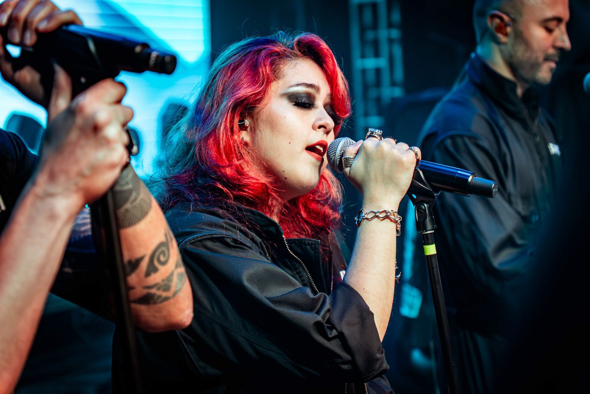 Uma artista com cabelos ruivos vibrantes cantando em um microfone no palco, com um músico parcialmente visível ao fundo.