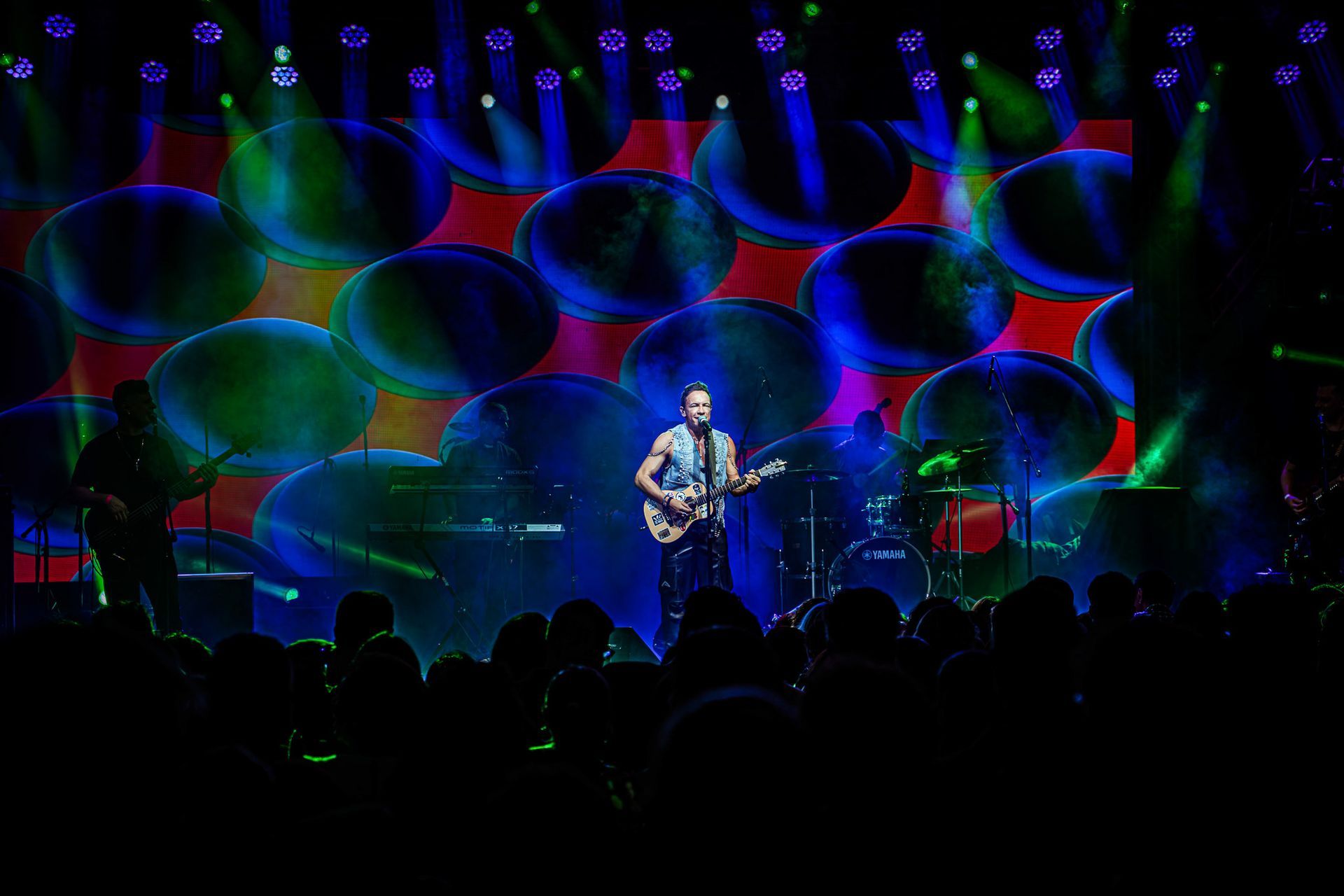Um músico toca guitarra em um palco iluminado por holofotes azuis e verdes contra um fundo de grandes bolhas escuras.