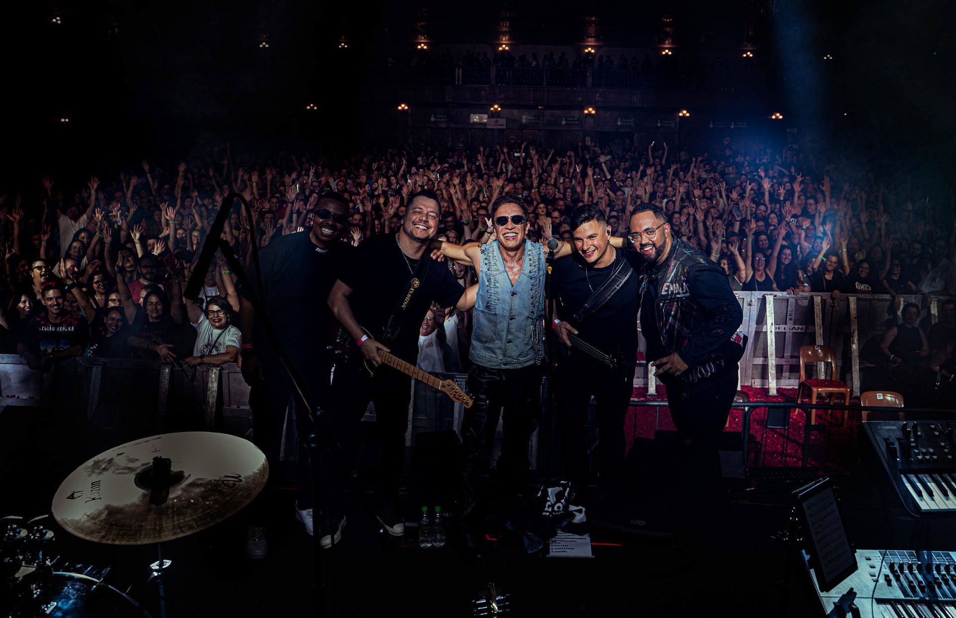 Uma banda posa no palco em frente a uma multidão entusiasmada durante um concerto, iluminada por luzes de espetáculo.