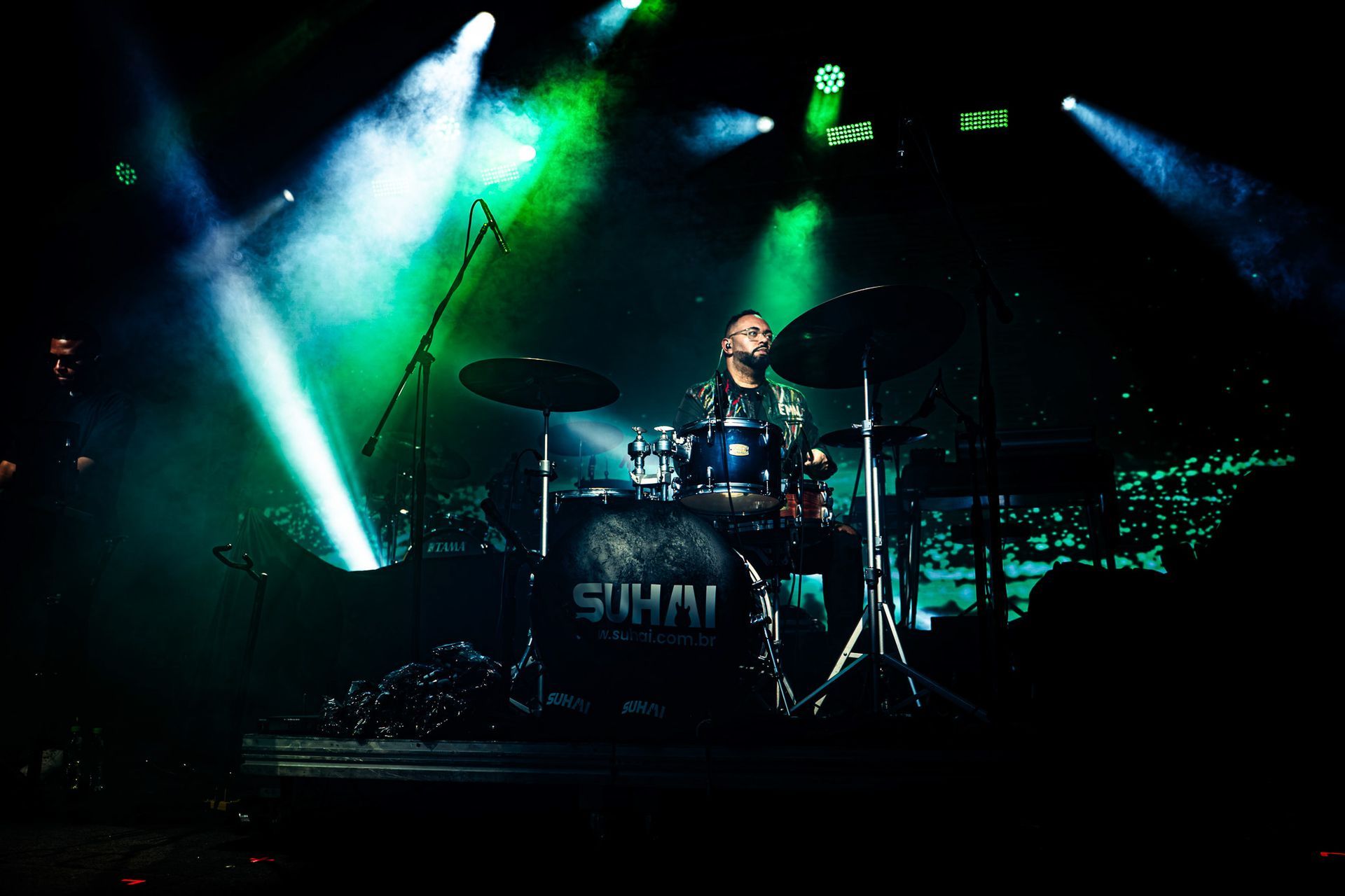 Baterista no palco, banhado por luzes verdes e azuis. A bateria exibe o nome 