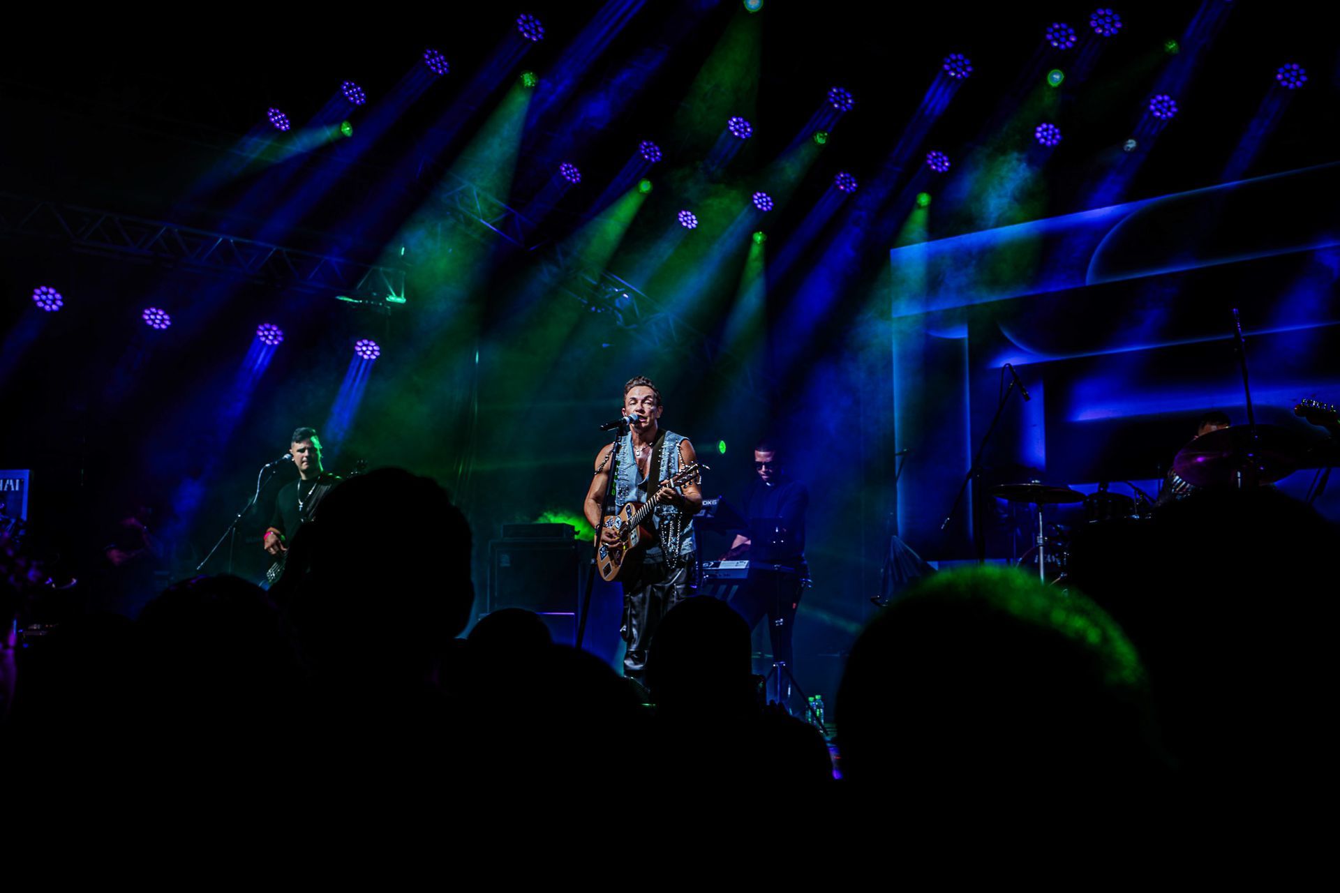 Um músico toca violão em um palco de concertos iluminado por holofotes vibrantes em tons de azul e verde.