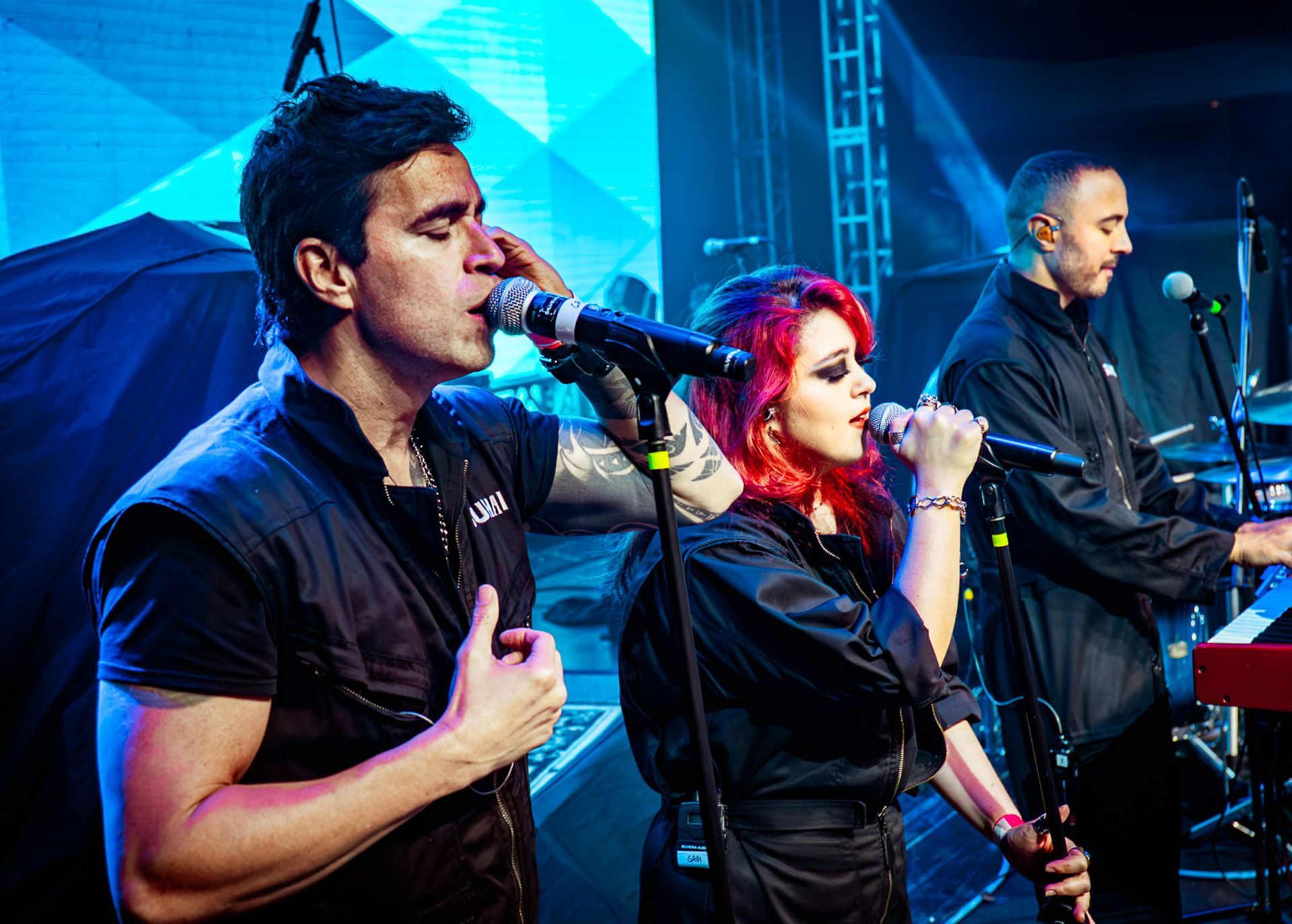 Dois cantores se apresentam em um palco com um tecladista, iluminados por luzes de palco azuis brilhantes.