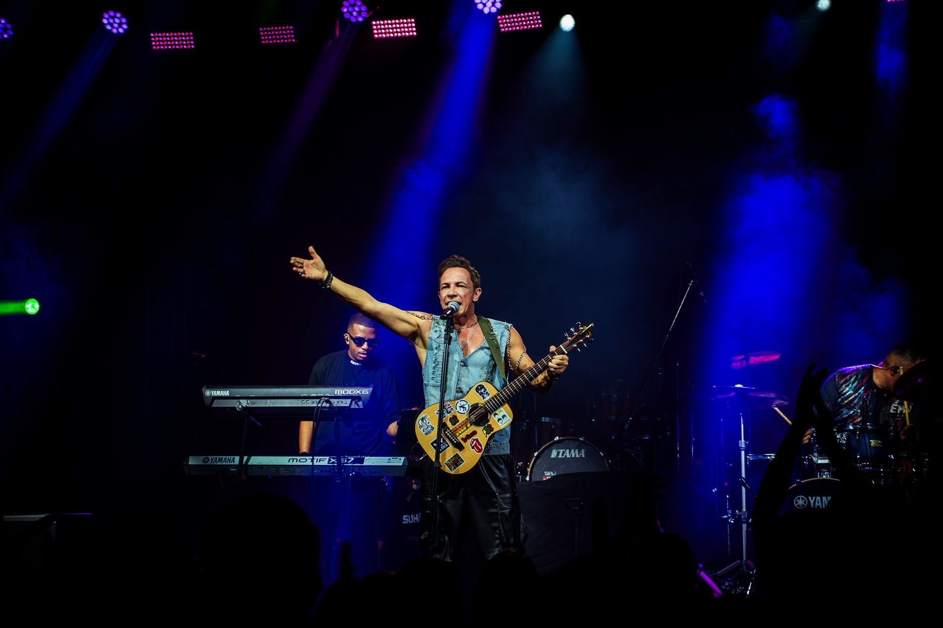 Um músico com um violão gesticula para a plateia sob luzes azuis de palco, com um tecladista ao fundo.