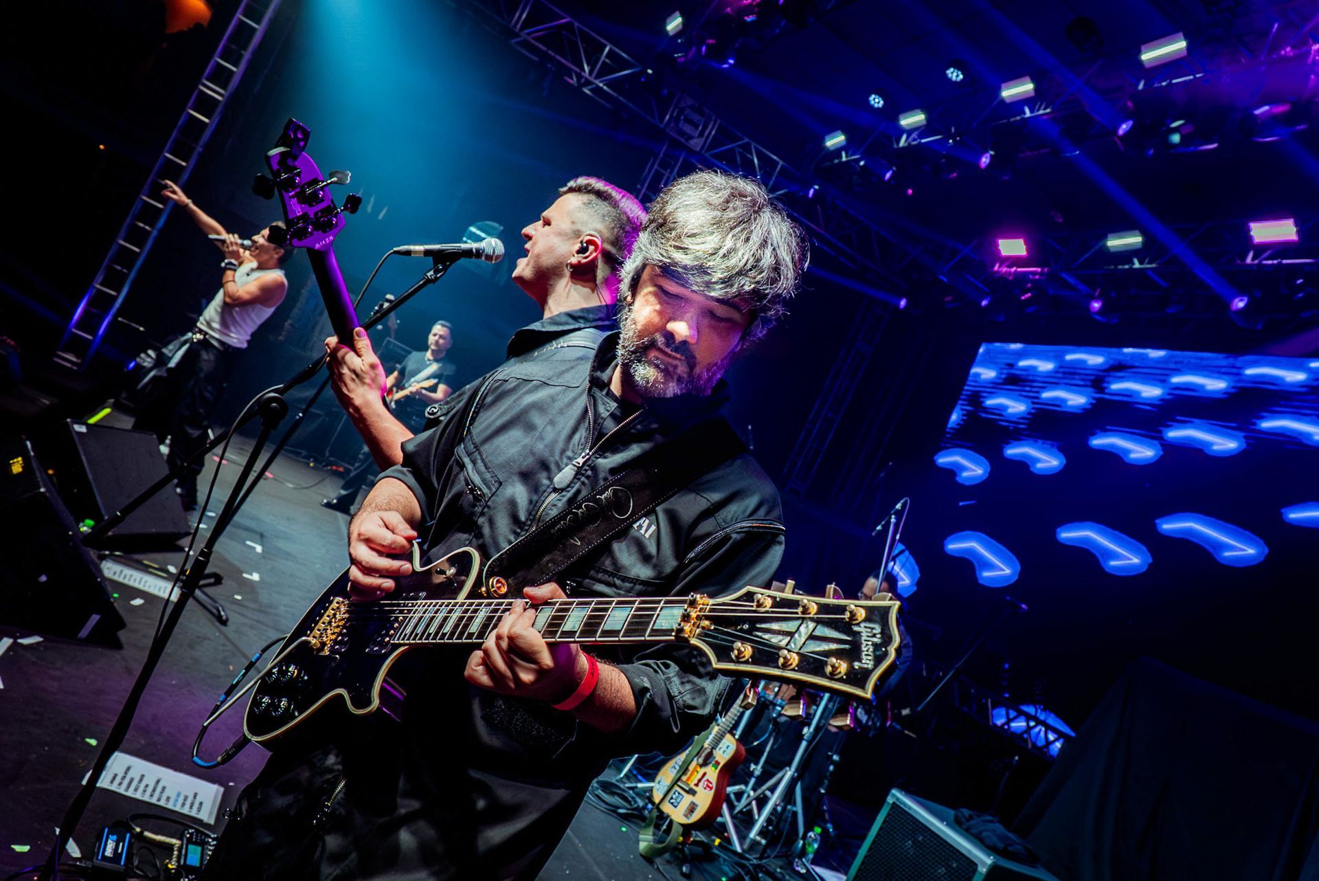 Um músico de cabelos grisalhos toca uma guitarra elétrica em um palco com iluminação azul, enquanto outros se apresentam.