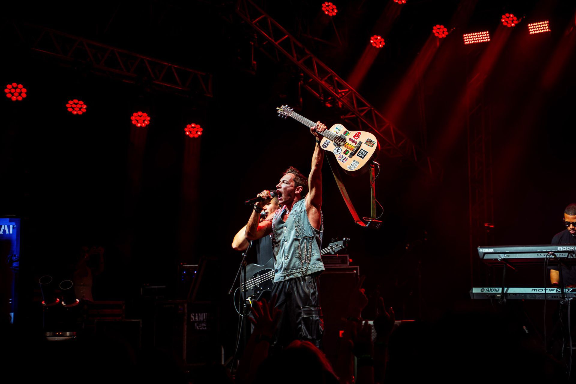 Um músico canta ao microfone enquanto ergue uma guitarra no palco, banhado por holofotes vermelhos.