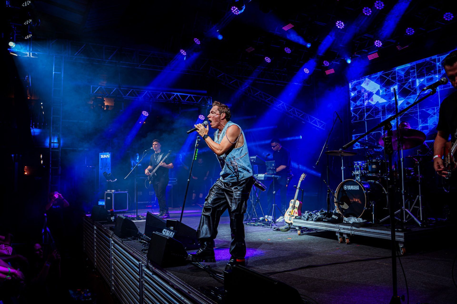 Um músico canta em um microfone em um palco iluminado acompanhado por um baterista e um baixista.