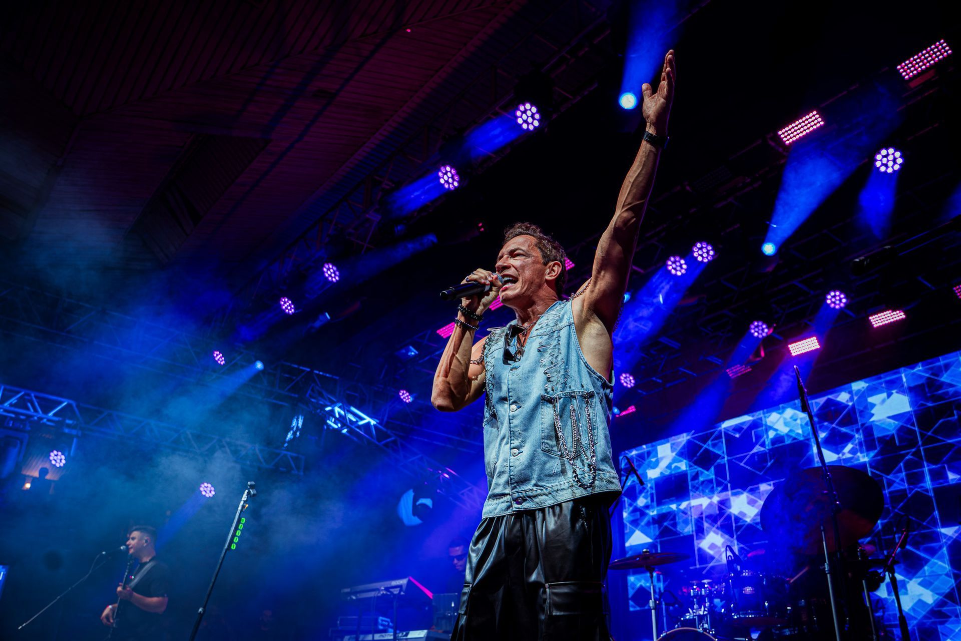 Um artista vestindo um colete jeans canta em um microfone com um braço levantado 