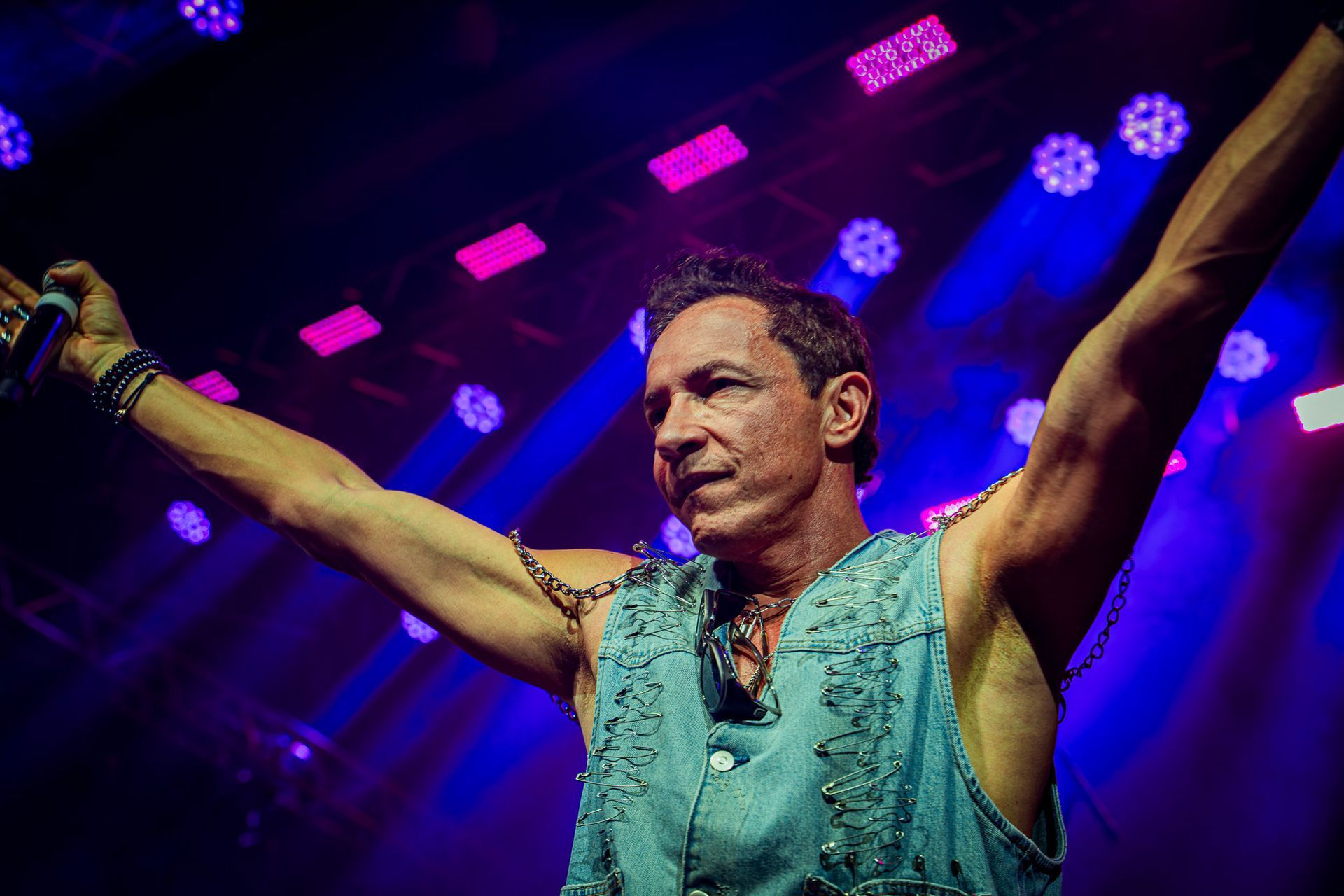 Um artista vestindo um colete jeans sem mangas está no palco com os braços estendidos sob luzes azuis brilhantes de um show.