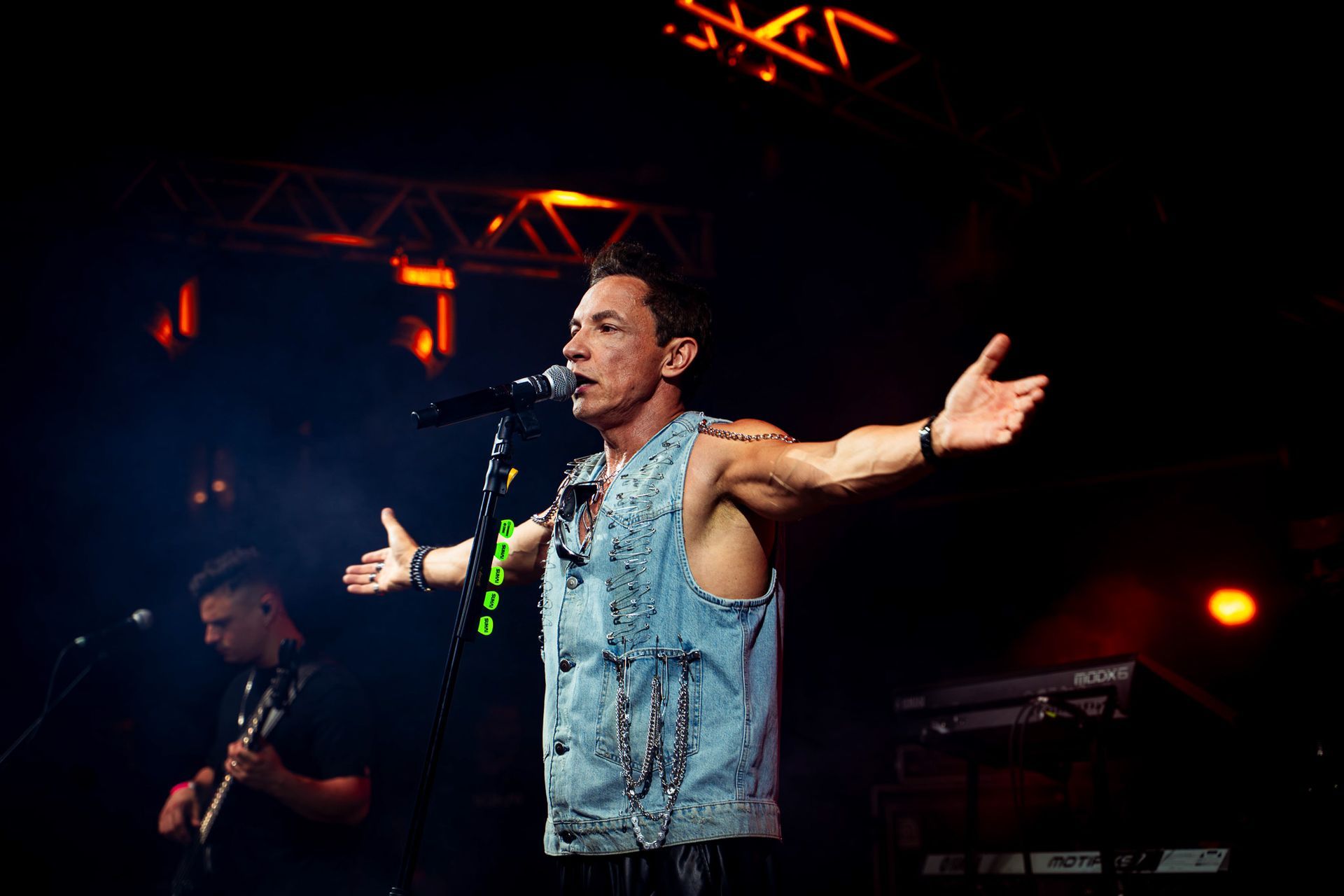 Um artista vestindo um colete jeans cravejado de tachas canta em um microfone com os braços abertos em um palco 