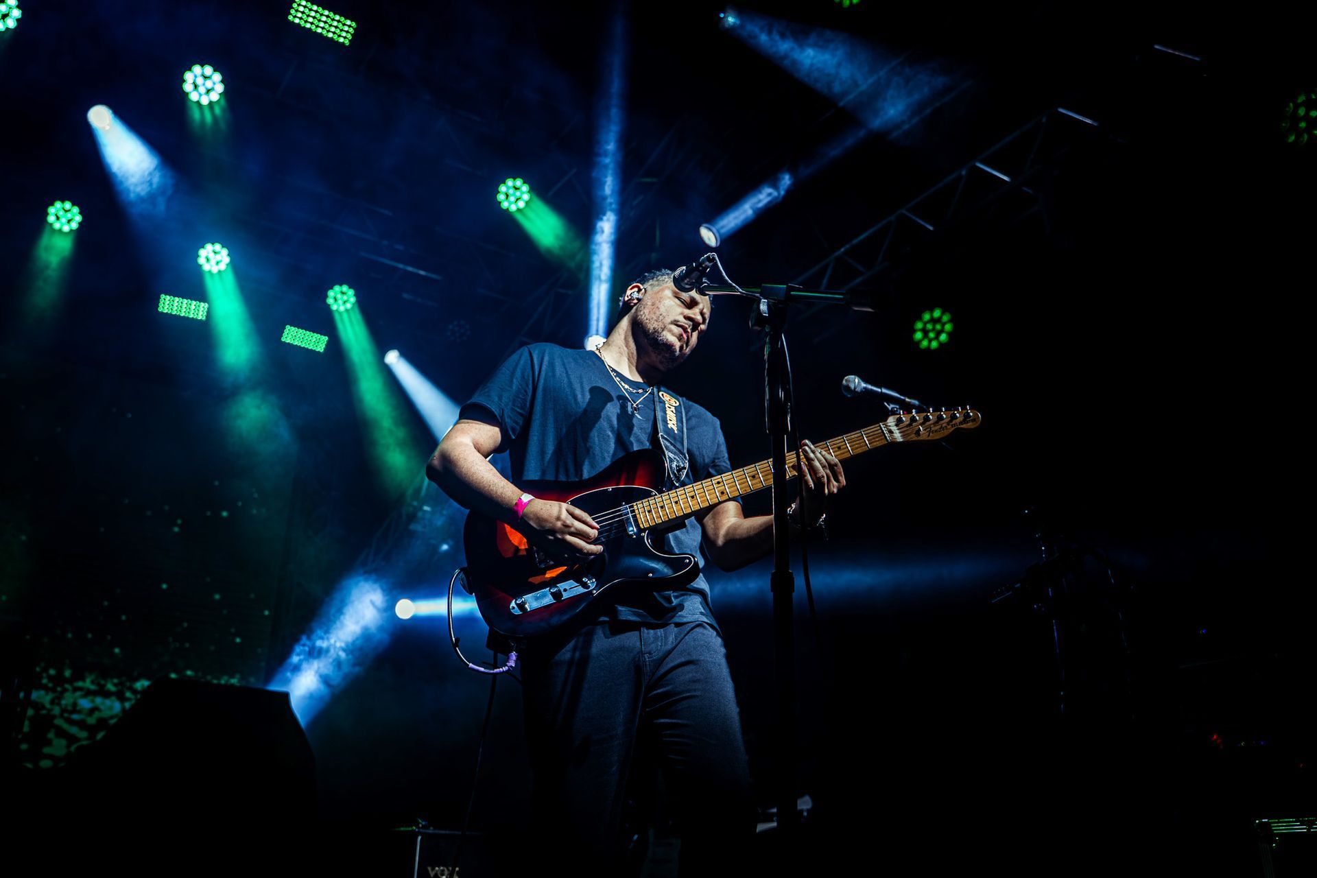 Guitarrista no palco, iluminado por holofotes verdes e azuis.