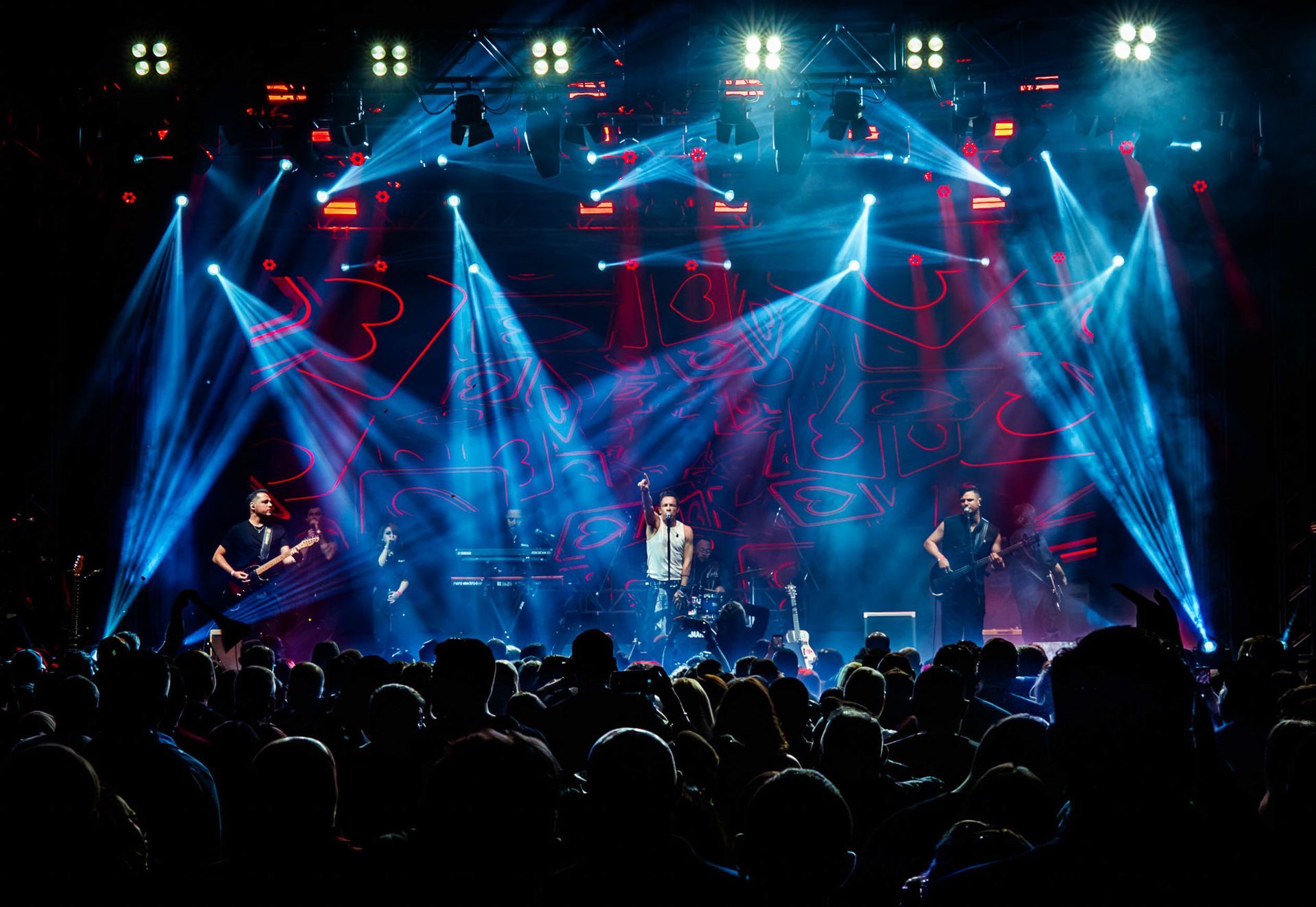 Uma banda se apresenta em um palco iluminado por holofotes azuis brilhantes,  diante de uma grande plateia.
