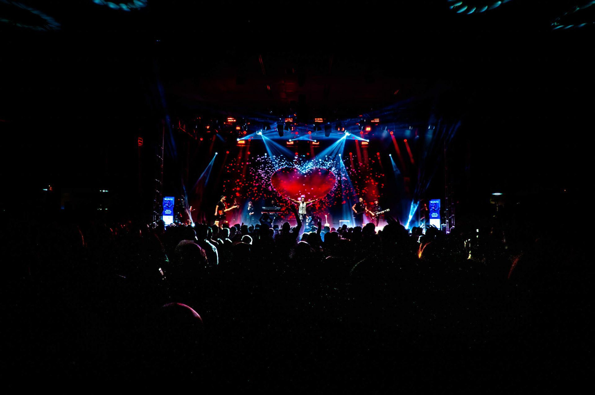Um palco de concertos iluminado por holofotes, com um artista no centro do palco e uma plateia escura
