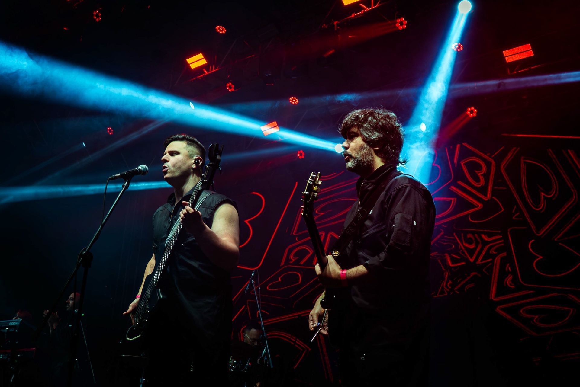 Dois músicos se apresentam no palco sob holofotes azuis, com um fundo composto por corações de néon vermelho.