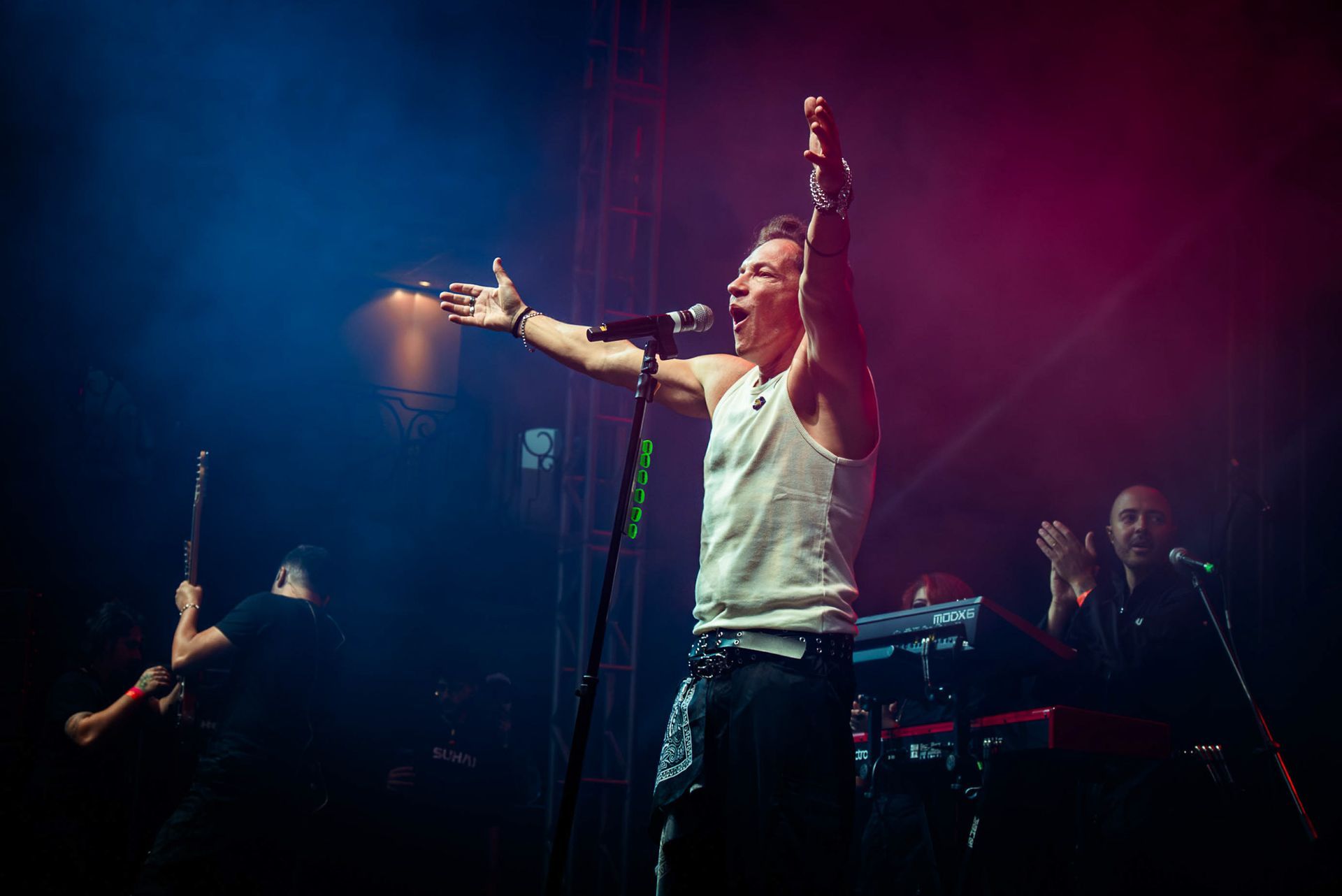 Um cantor de regata branca se apresenta no palco com os braços erguidos, banhado por luzes de concerto azuis e vermelhas.
