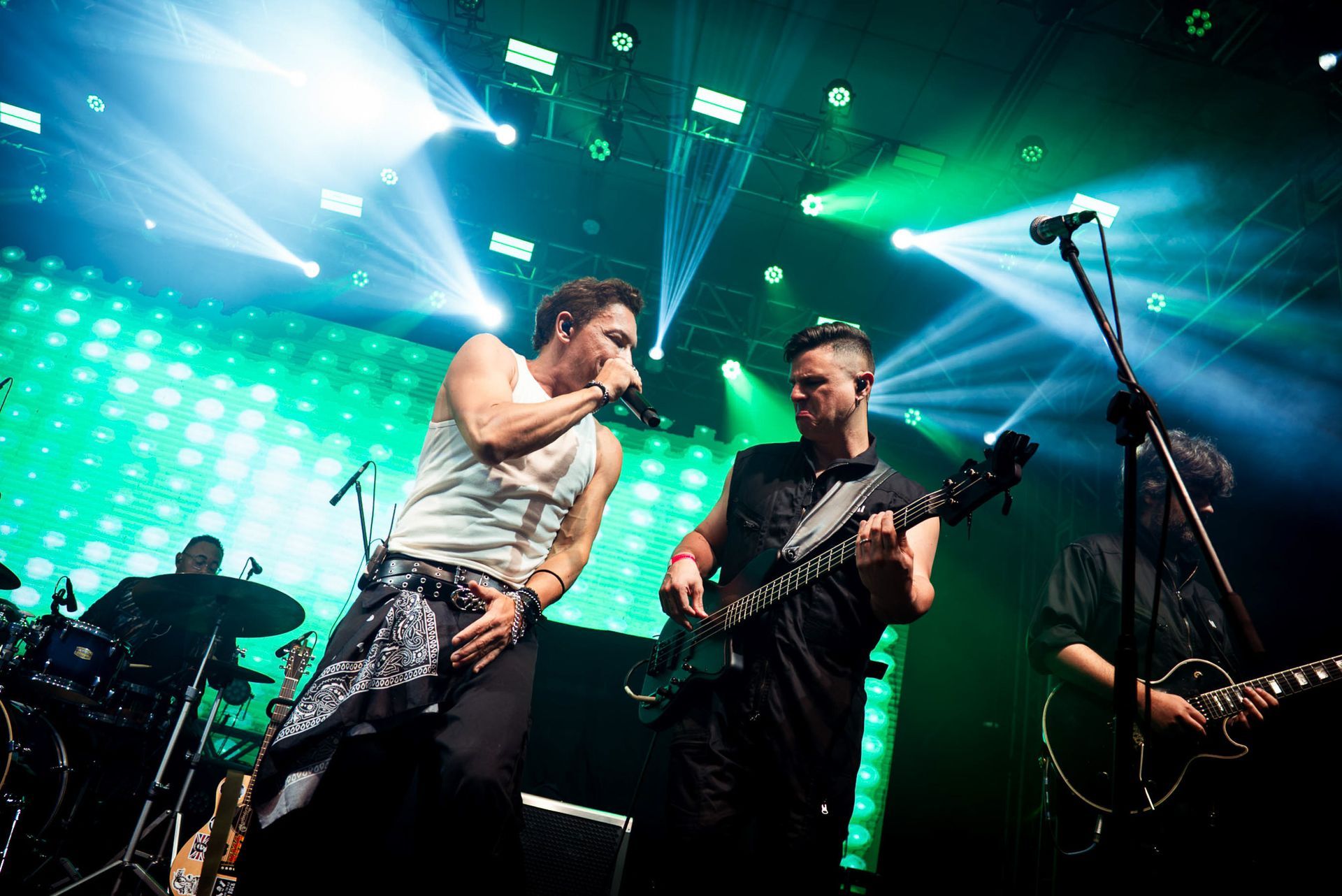 Uma banda se apresenta no palco sob holofotes com um fundo colorido e estampado, enquanto o vocalista segura um microfone.