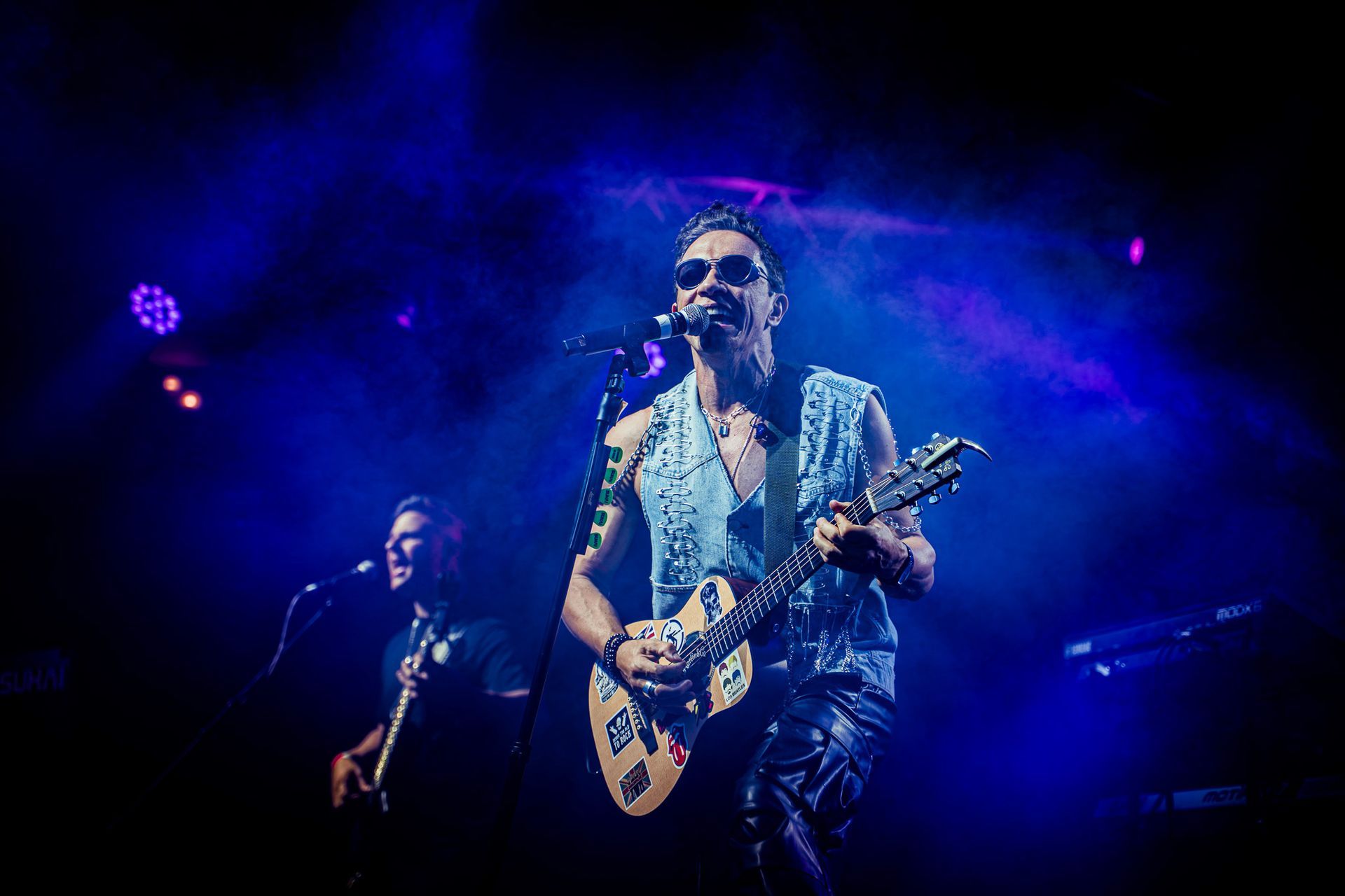 Dois músicos no palco sob luzes azuis; um deles canta ao microfone enquanto toca guitarra.
