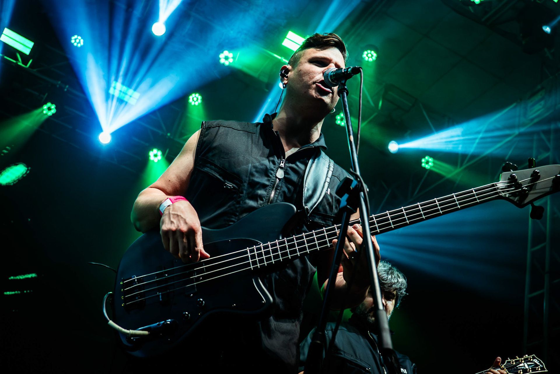 Um músico cantando ao microfone enquanto toca um baixo elétrico escuro em um palco iluminado por holofotes azuis e verdes.