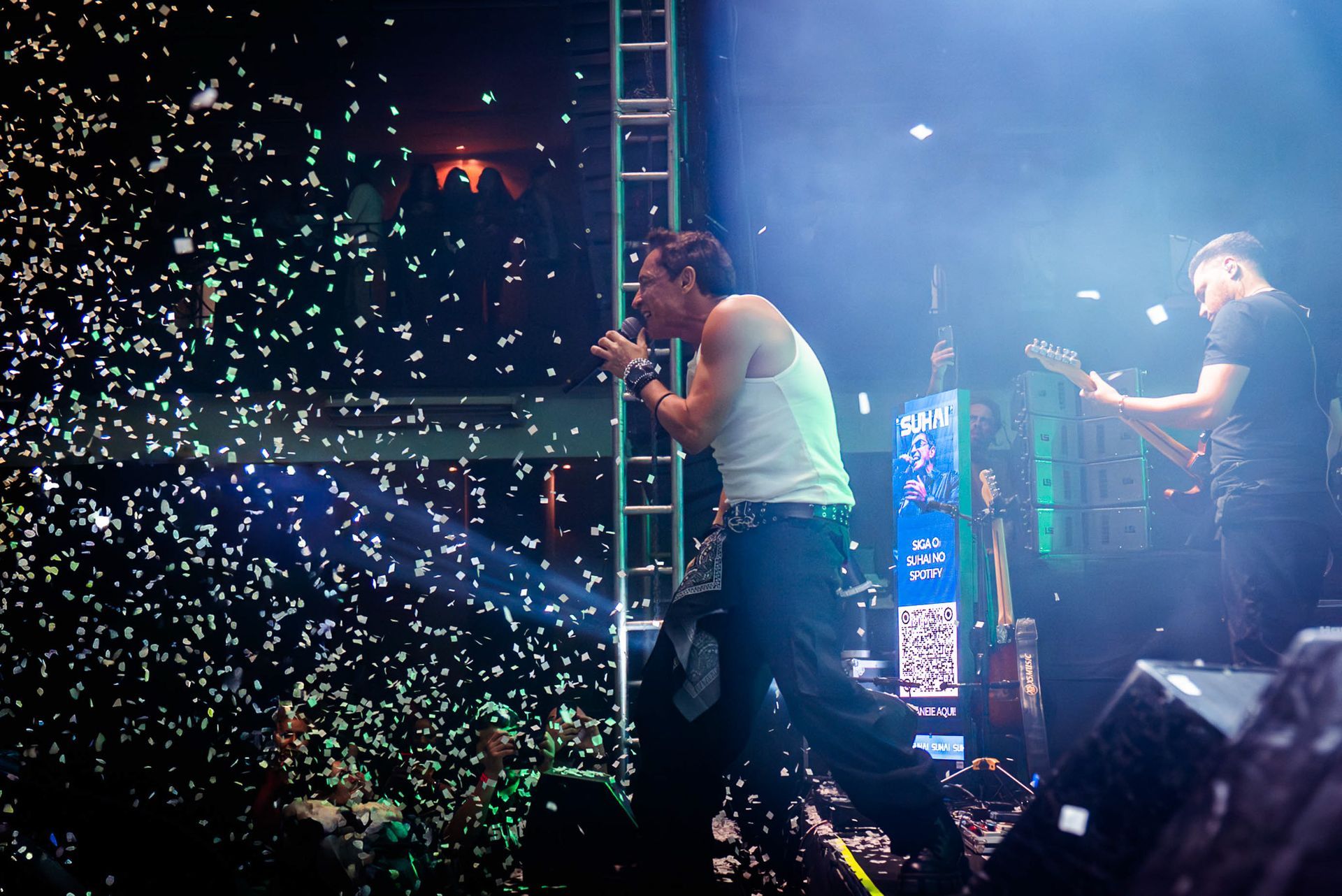 Um músico canta em um microfone no palco em meio a uma chuva de confetes, enquanto outra pessoa toca violão à direita.