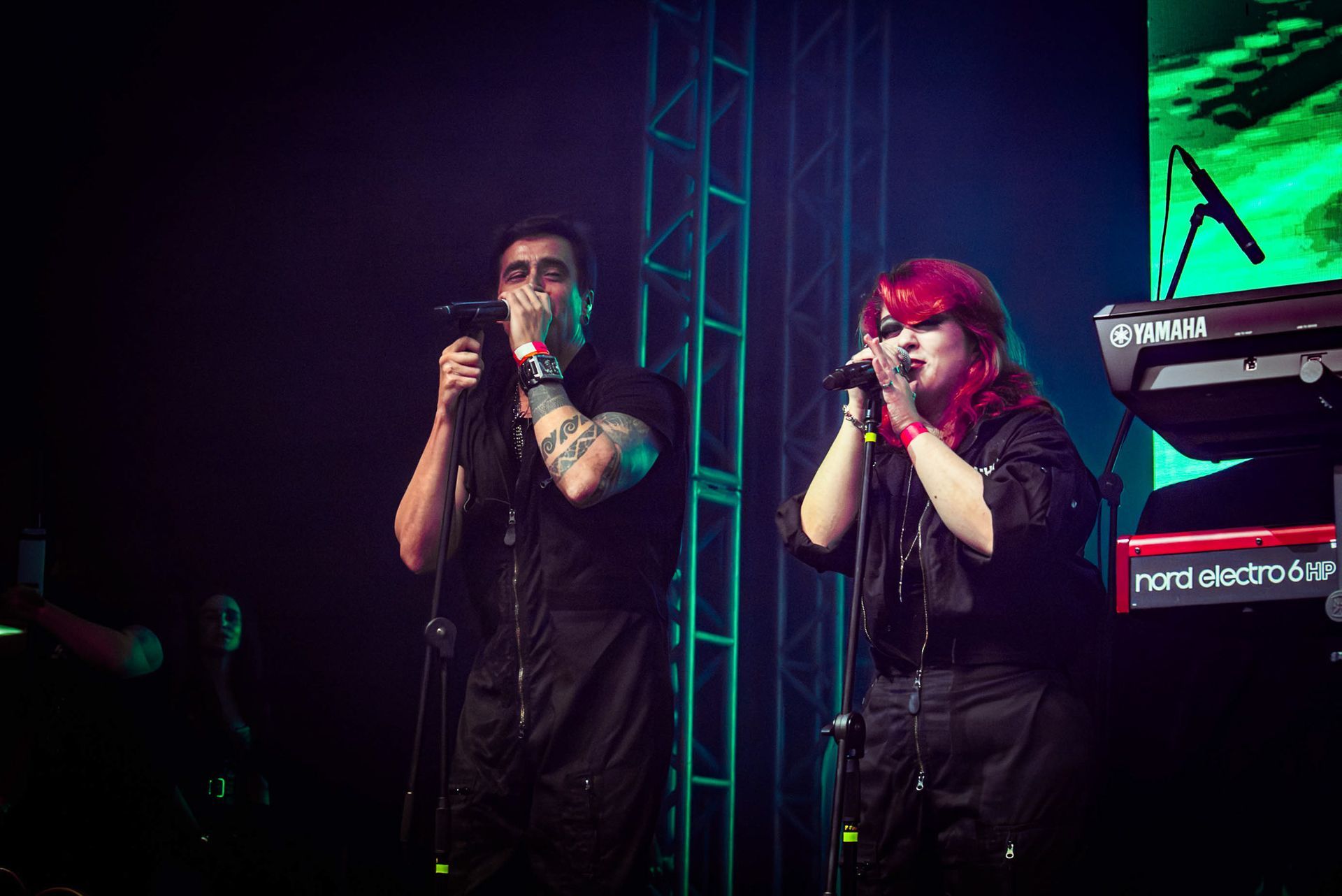 Vocalistas vestidos com macacões pretos cantam em microfones num palco com um teclado e um fundo de tela verde.
