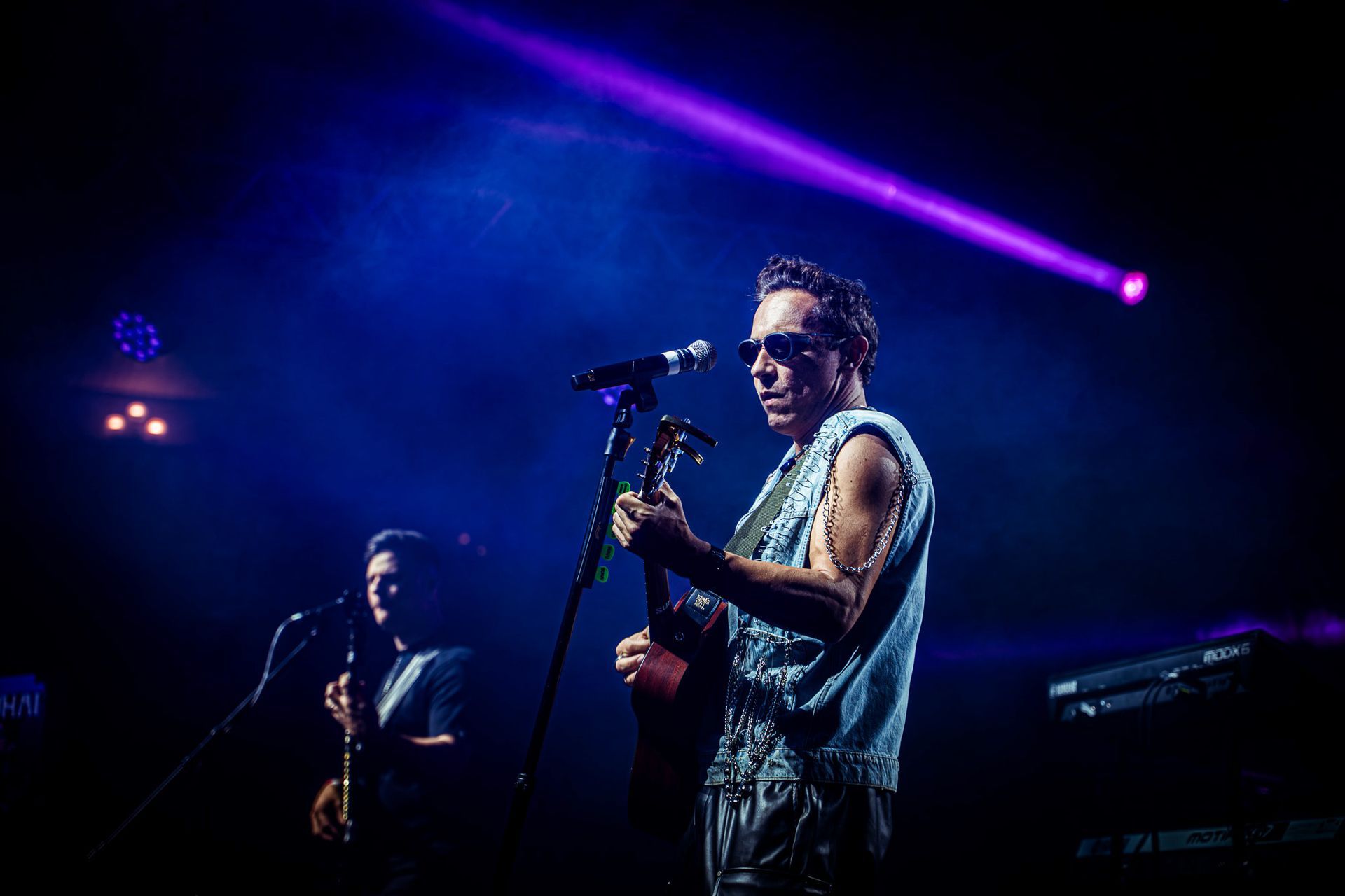 Um músico toca violão no palco sob um forte holofote roxo, com um colega de banda visível ao fundo.