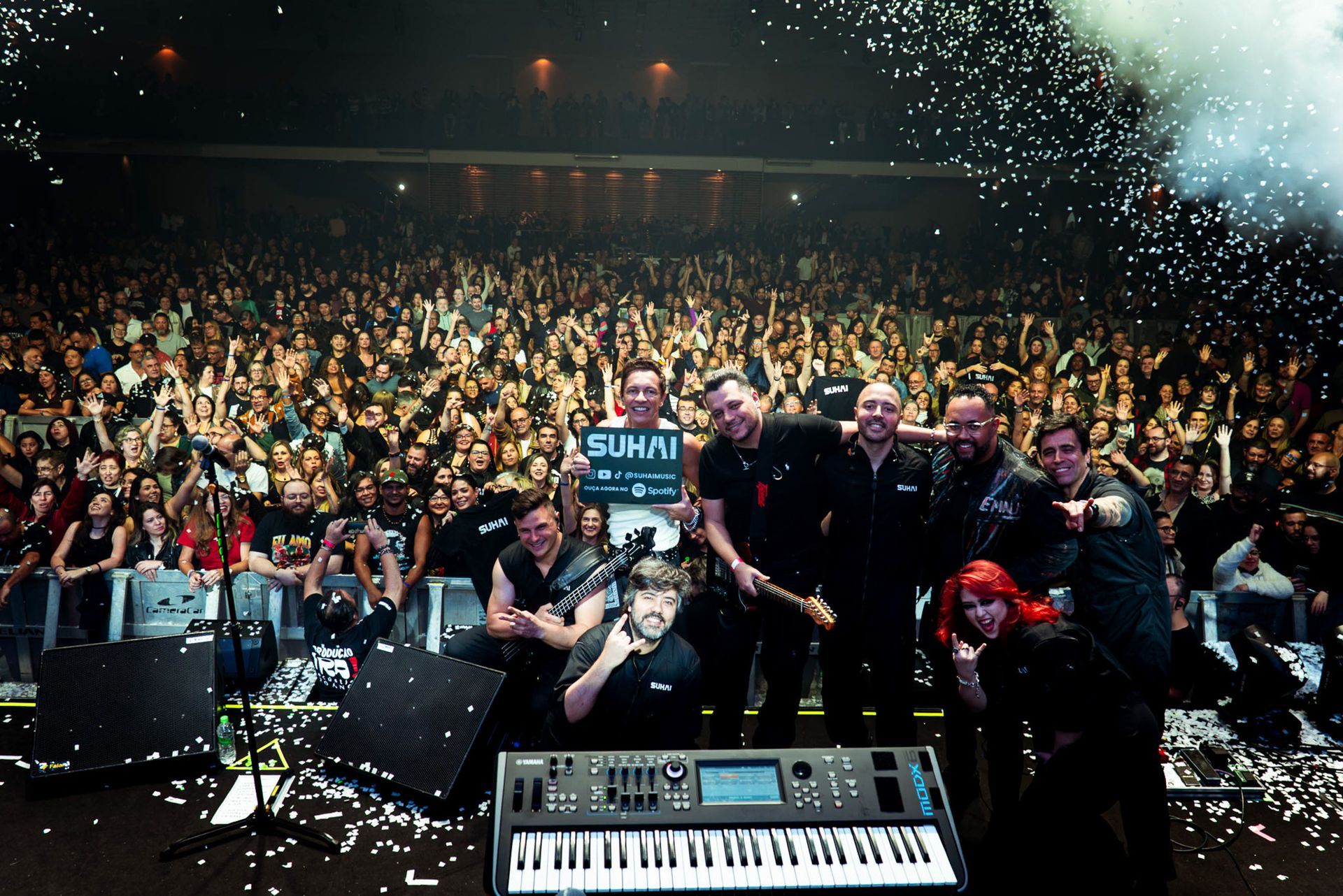 Uma banda posa para uma foto no palco em frente a uma grande multidão entusiasmada durante um show com confetes no ar.
