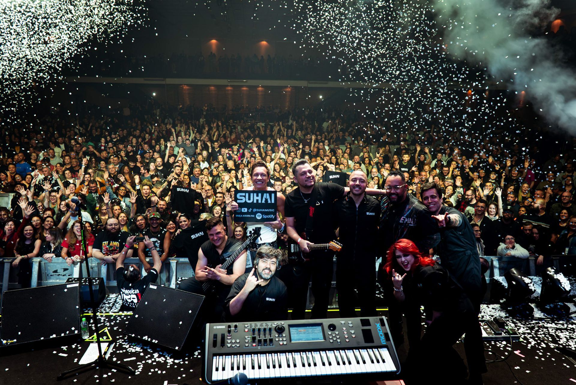 Um grupo musical está no palco diante de uma grande multidão que os aplaude, enquanto confetes caem ao seu redor.