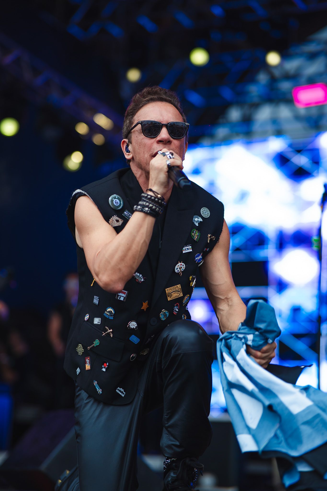 Um artista de óculos escuros cantando, ajoelhado no palco, vestindo um colete com broches e segurando uma bandeira azul e branca.