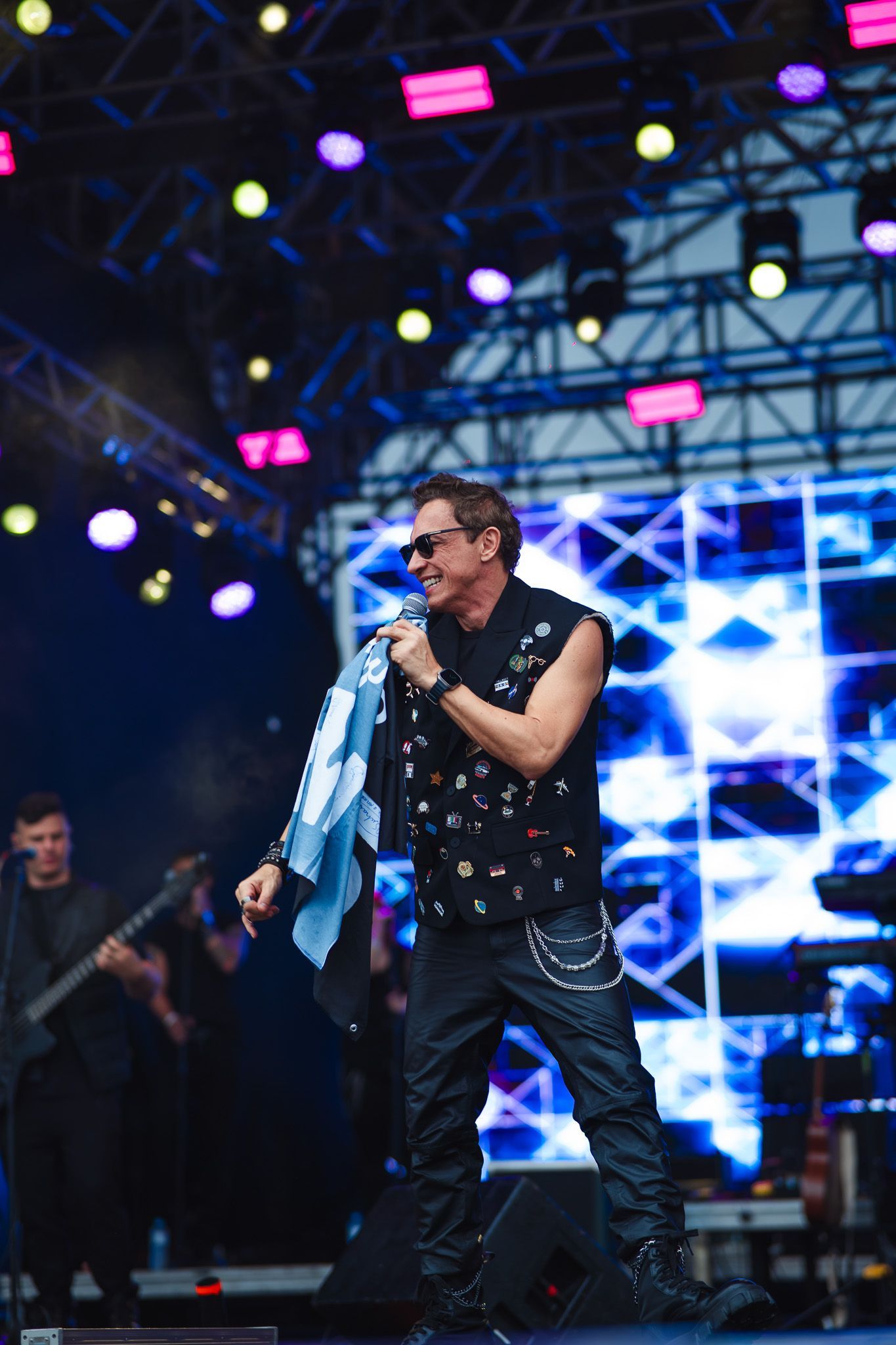 Um artista cantando no palco, usando óculos escuros e roupas pretas, segurando uma toalha. Cenário de show com luzes.