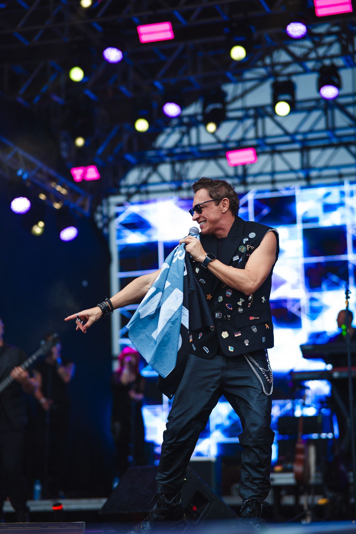 Um artista se apresentando no palco, apontando com o microfone na mão, usando óculos escuros e colete. Luzes de palco ao fundo.