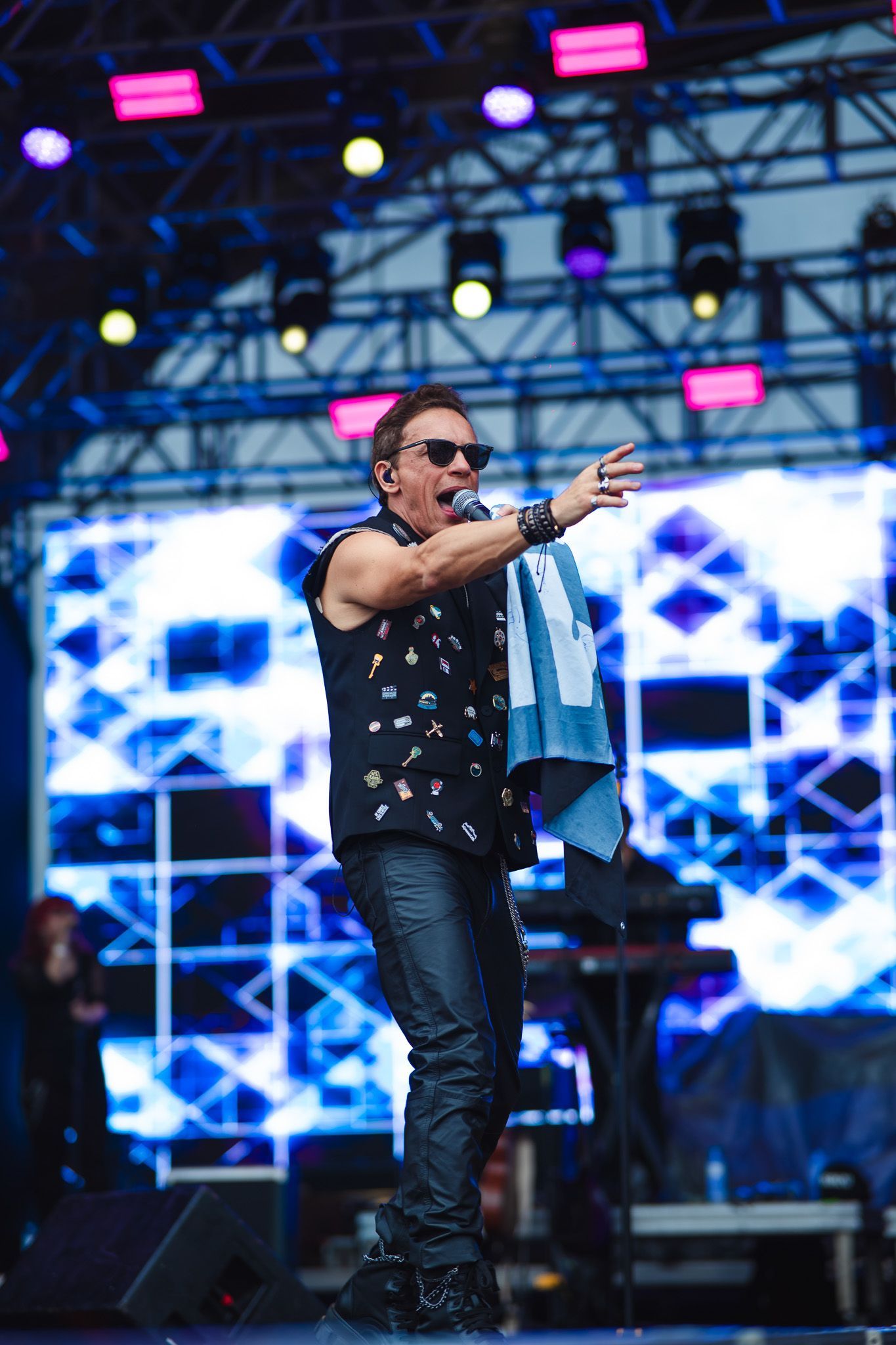 Um artista no palco com óculos escuros, apontando e segurando uma toalha. Palco iluminado com luzes azuis e rosas.