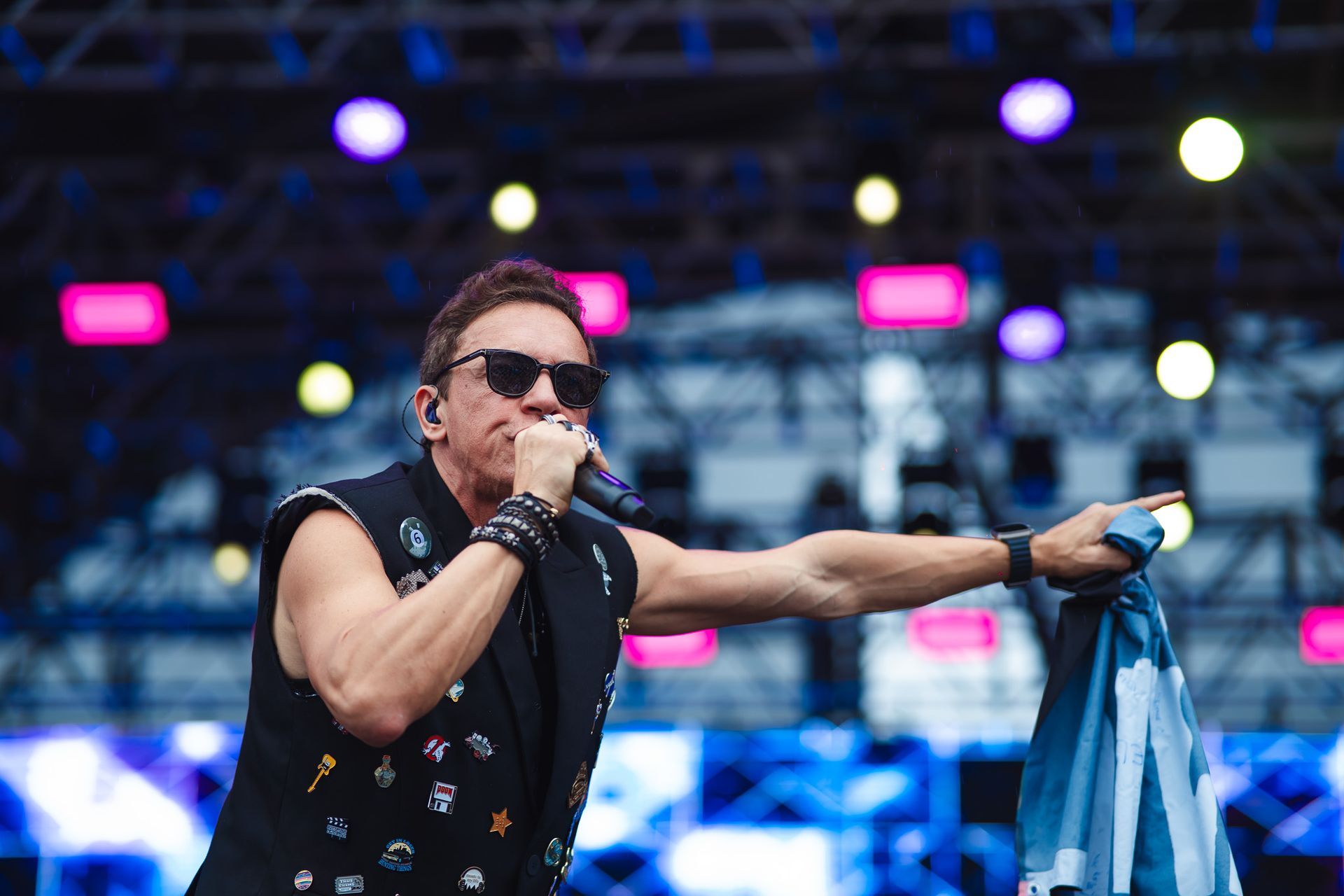 Um artista de óculos escuros cantando em um microfone no palco, apontando com a mão direita, segurando um pano azul.