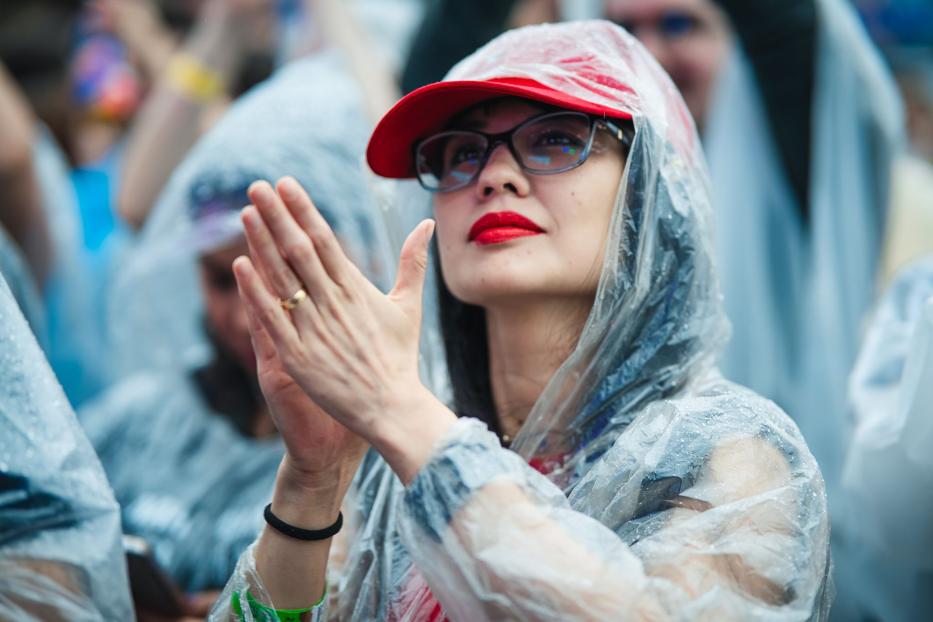 Mulher usando boné vermelho, óculos e capa de chuva bate palmas em um evento ao ar livre.