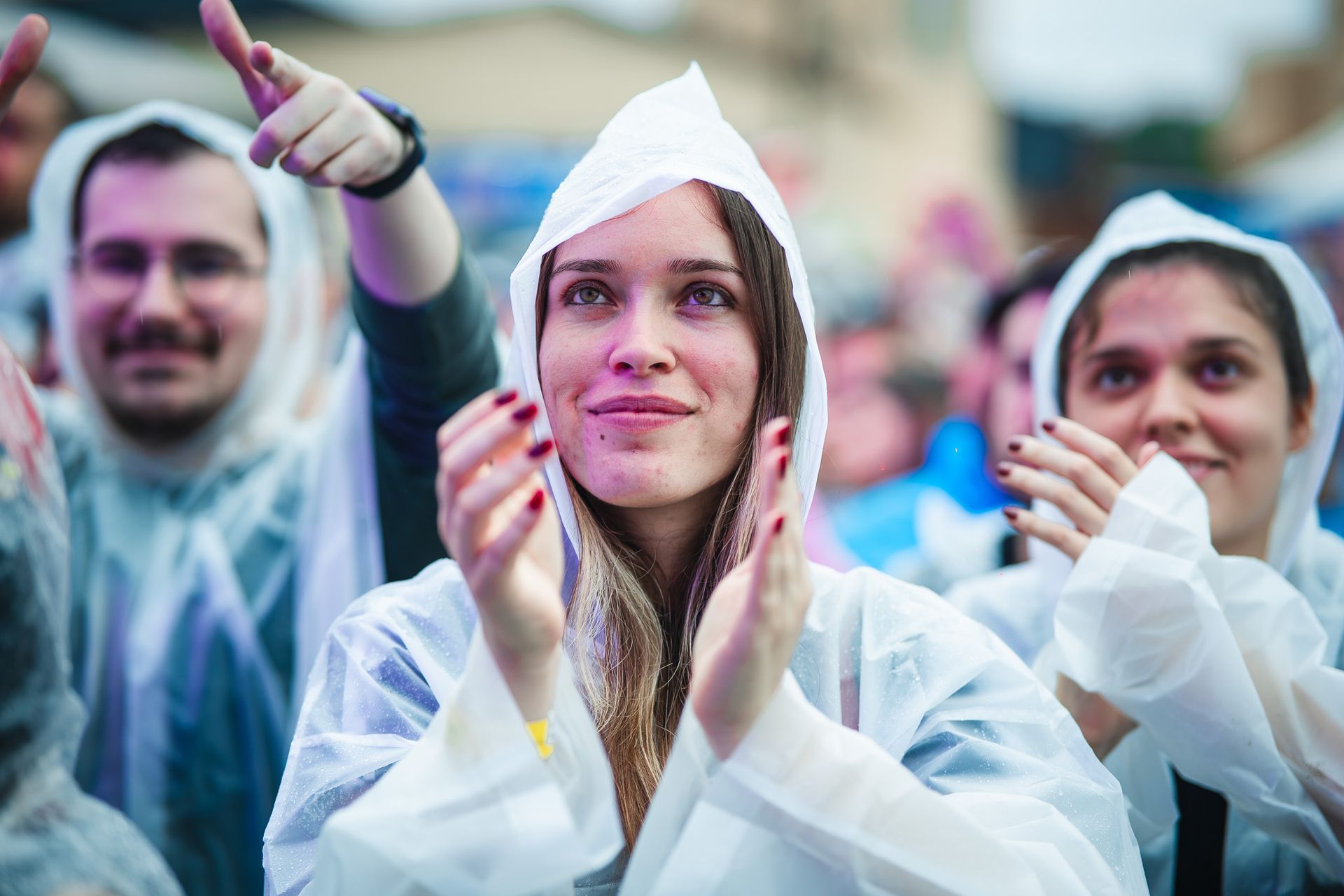 Pessoas de capa de chuva aplaudindo em um evento ao ar livre; sorrindo e concentradas no palco.