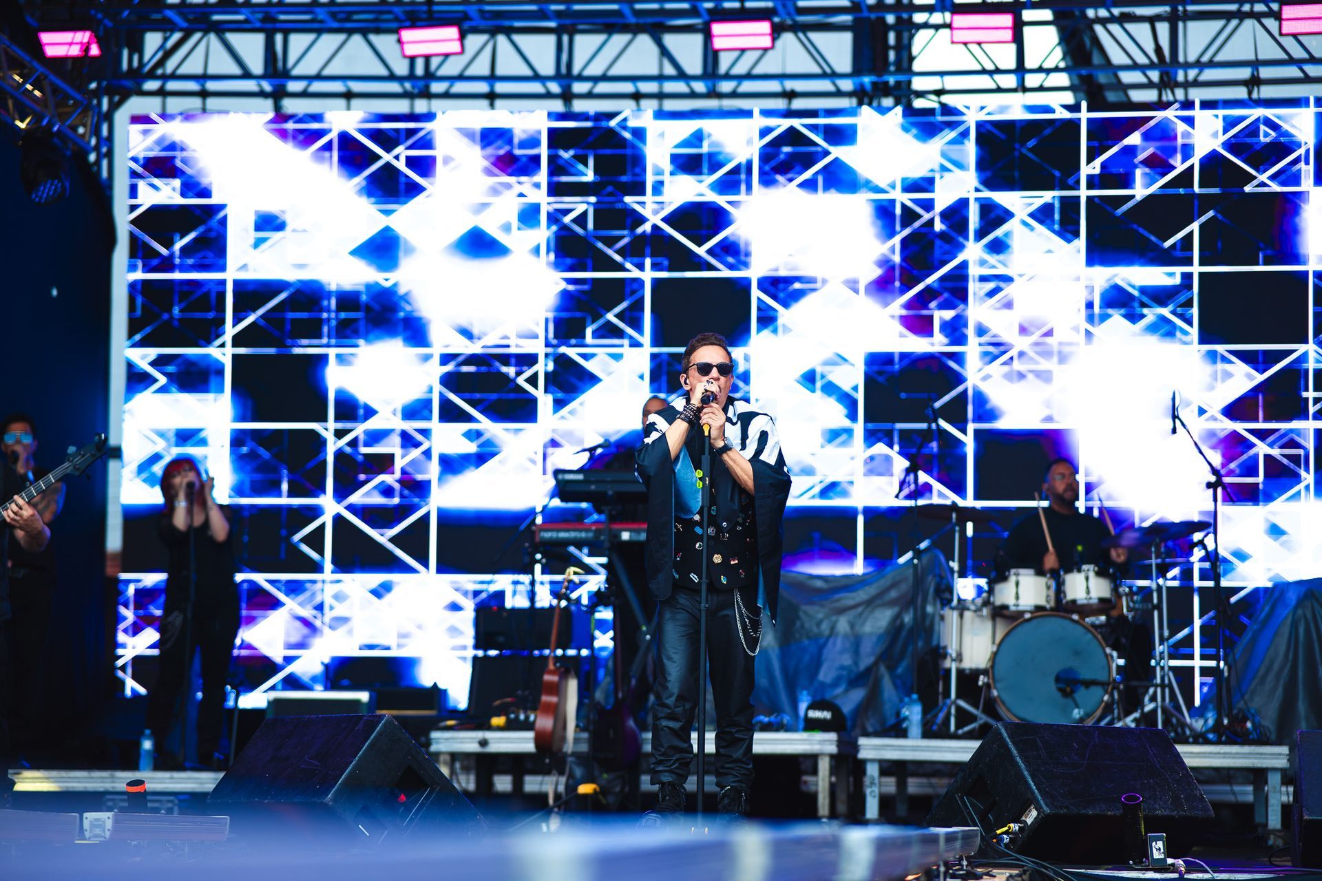 Banda se apresentando no palco com fundo iluminado em azul. Vocalista de óculos escuros, bateria, teclado, guitarra