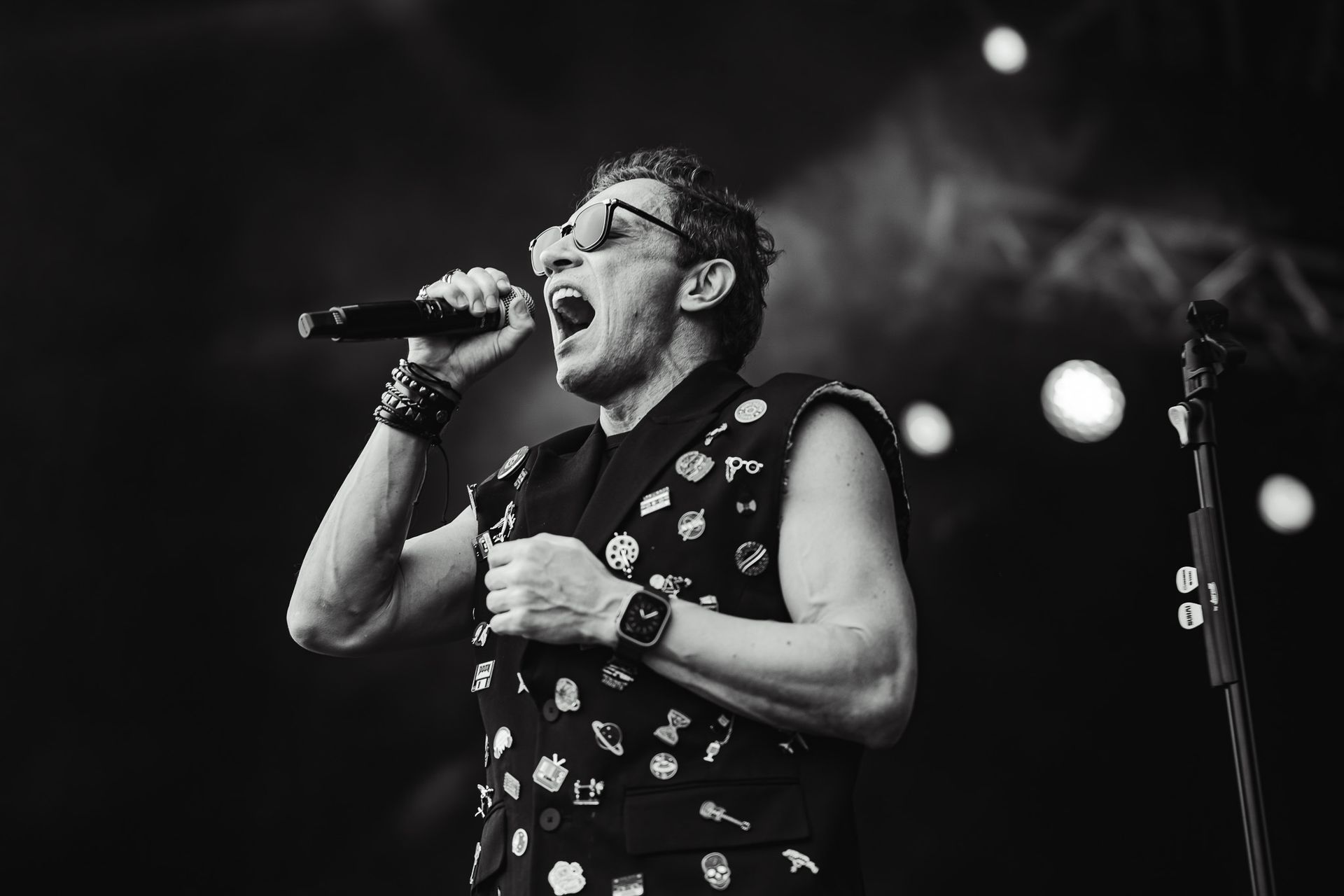 Um artista cantando ao microfone no palco, usando óculos escuros e colete. Foto em preto e branco.