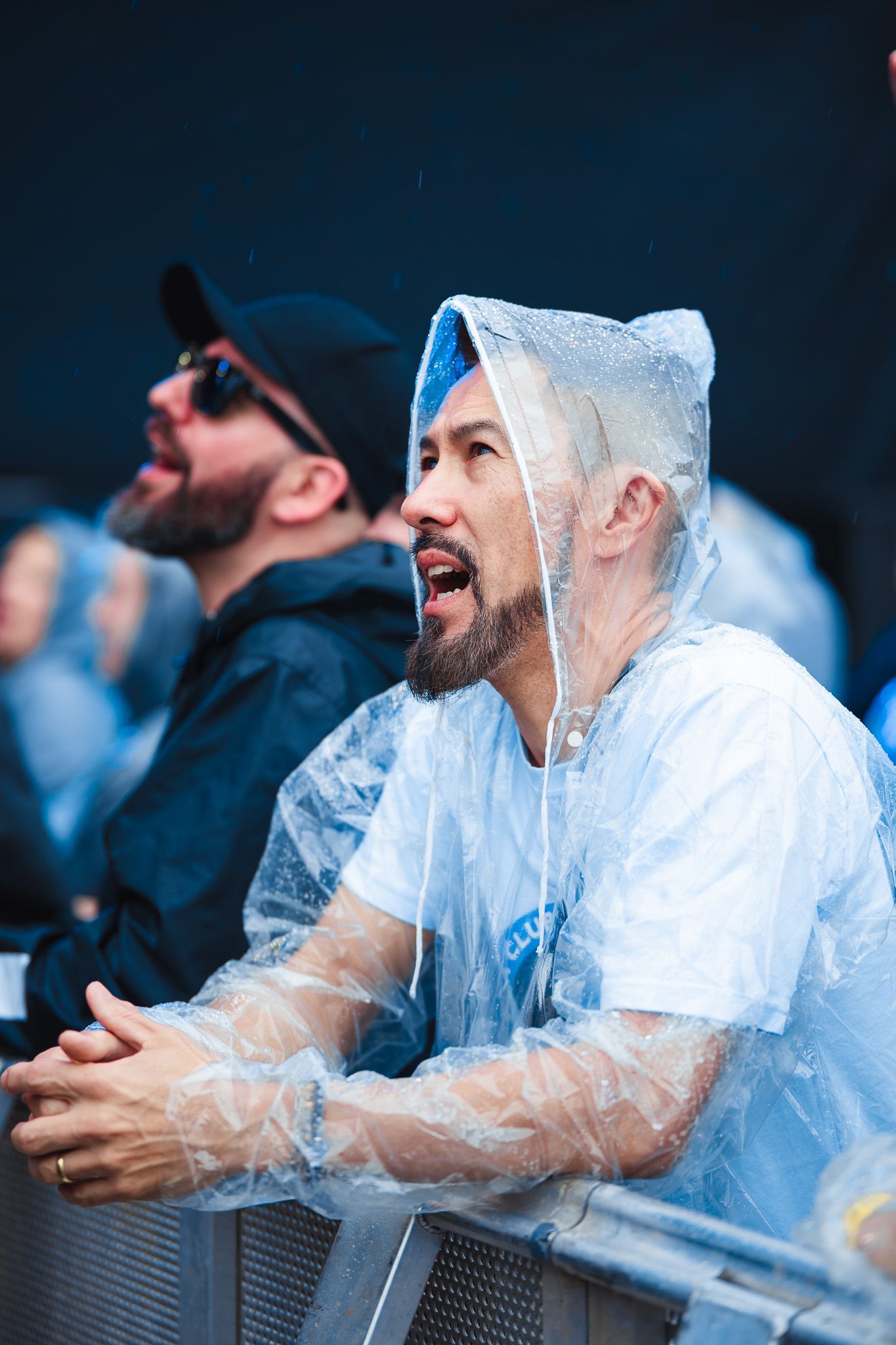 Um homem com capa de chuva transparente observa o evento. Outro homem, de chapéu preto e óculos escuros.