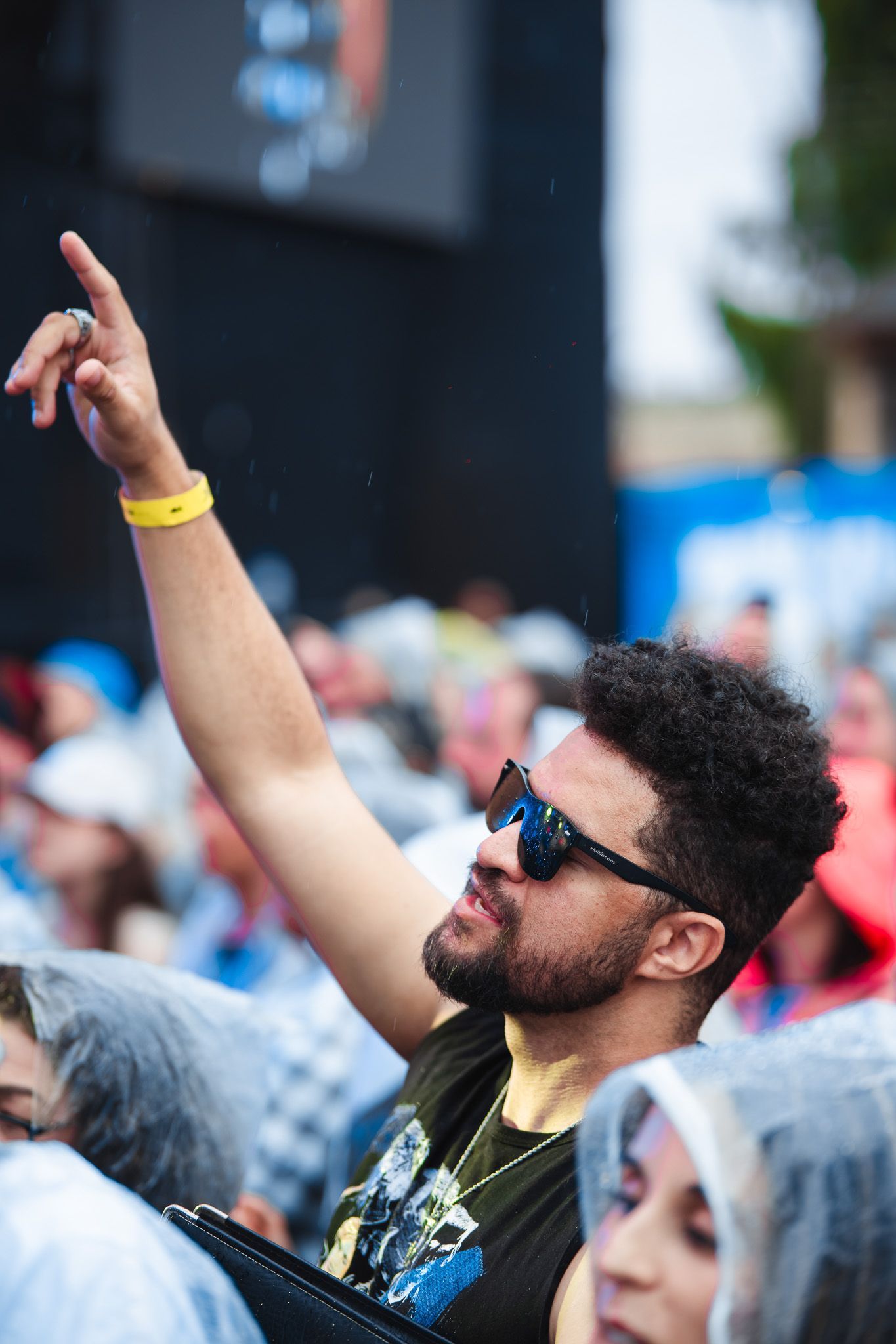Homem em um evento ao ar livre, com o braço levantado, usando óculos de sol e uma pulseira amarela.