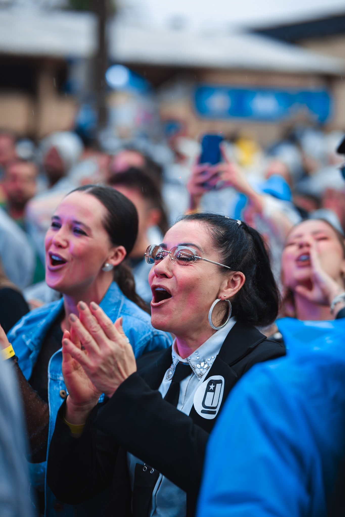 Pessoas vibrando em um evento ao ar livre, aplaudindo e gritando. Mulher de óculos e blazer preto ao centro.