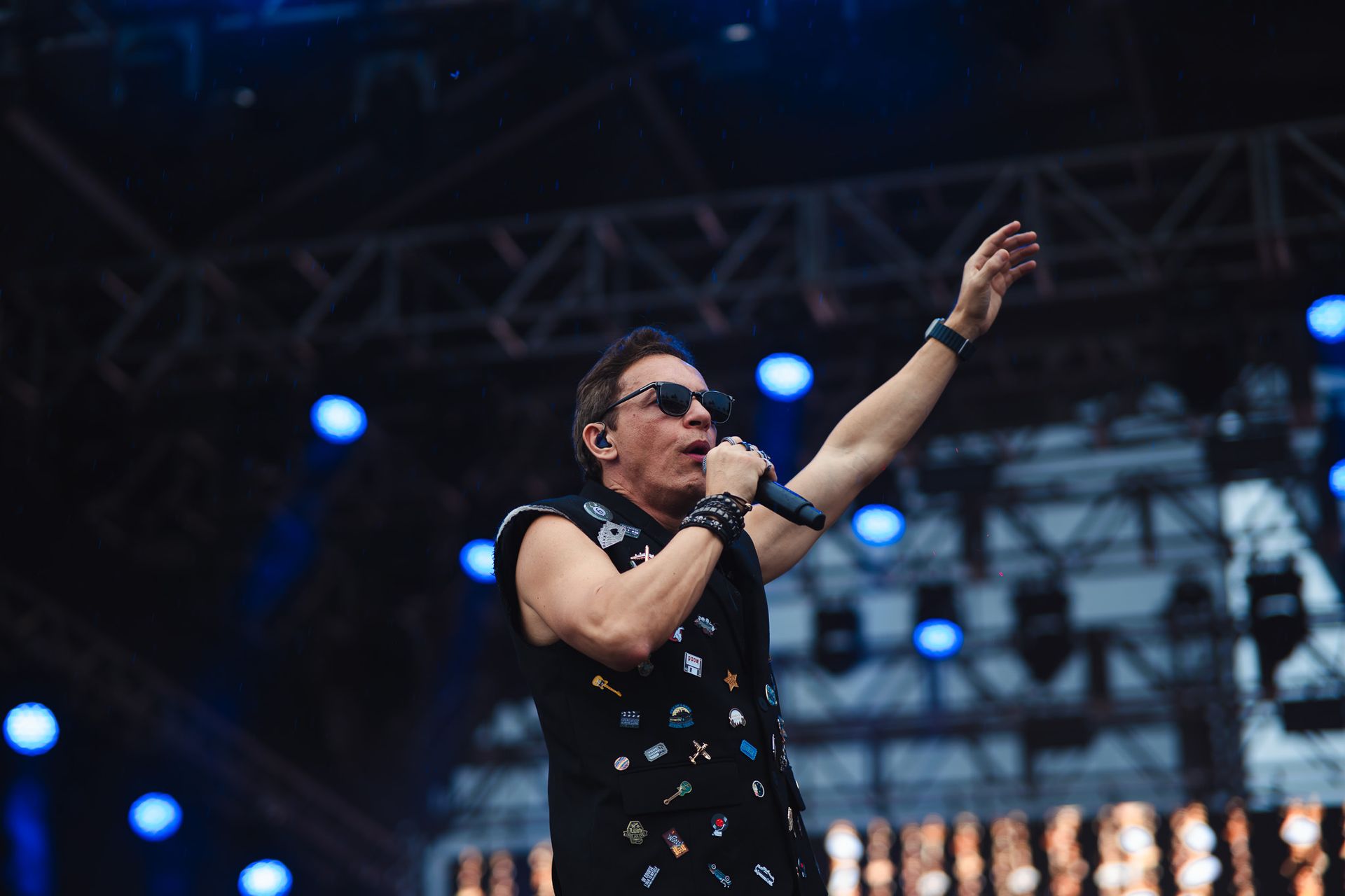 Um artista no palco cantando, com o braço levantado. Usando óculos escuros e uma camiseta preta sem mangas com broches.