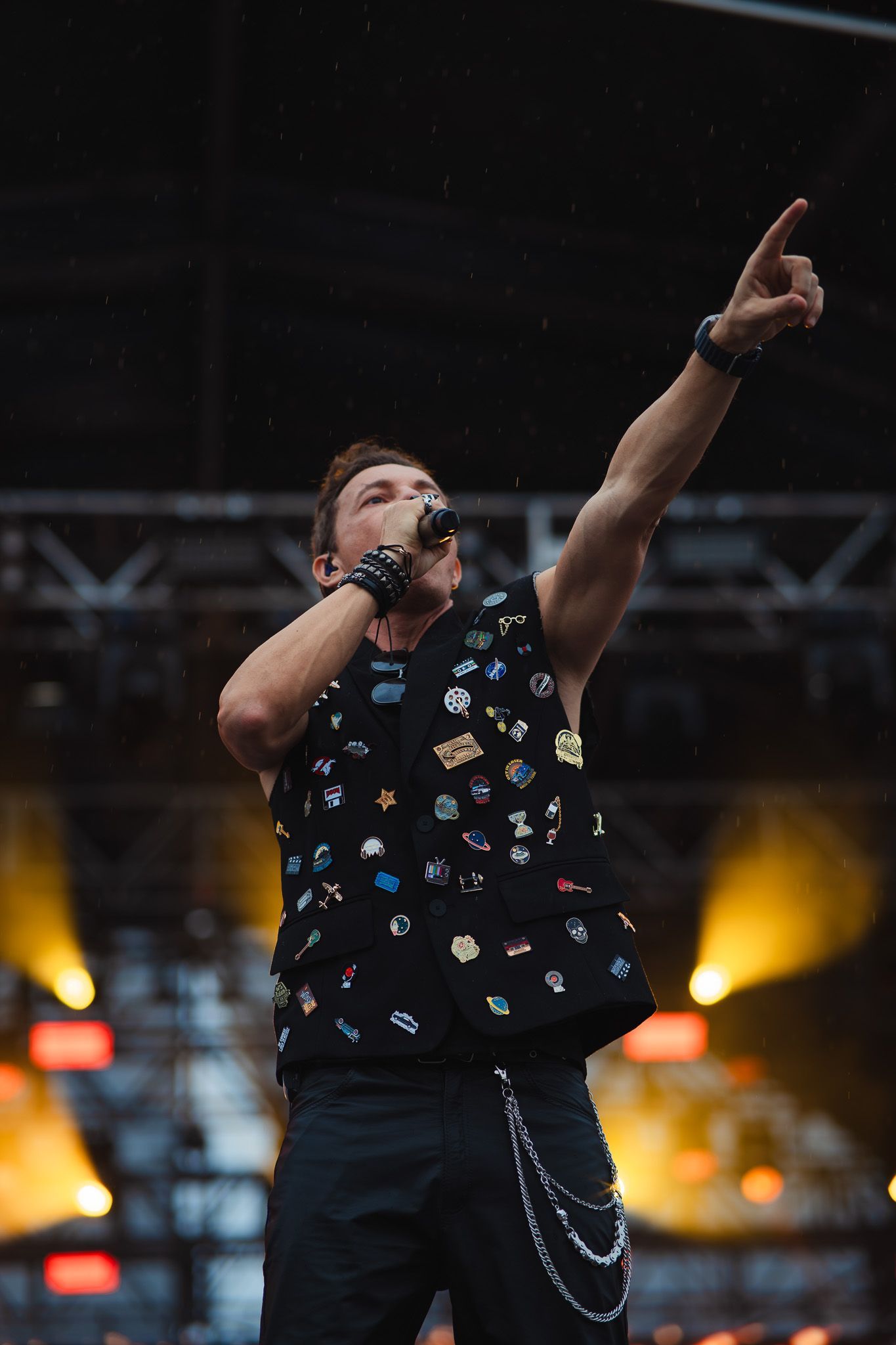 Um artista no palco, de colete coberto de alfinetes, cantando com um braço levantado e apontando.