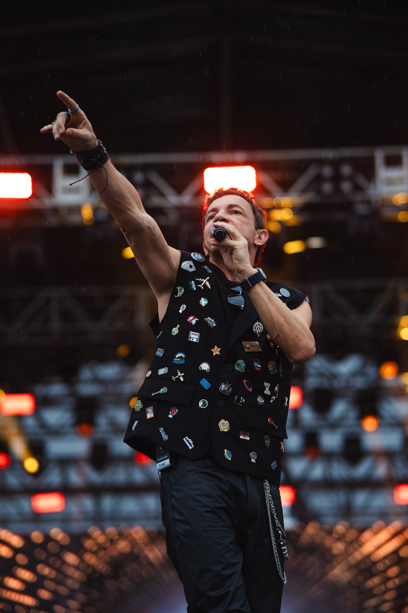 Um artista no palco cantando e apontando. Vestindo colete preto coberto de alfinetes, segurando um microfone.
