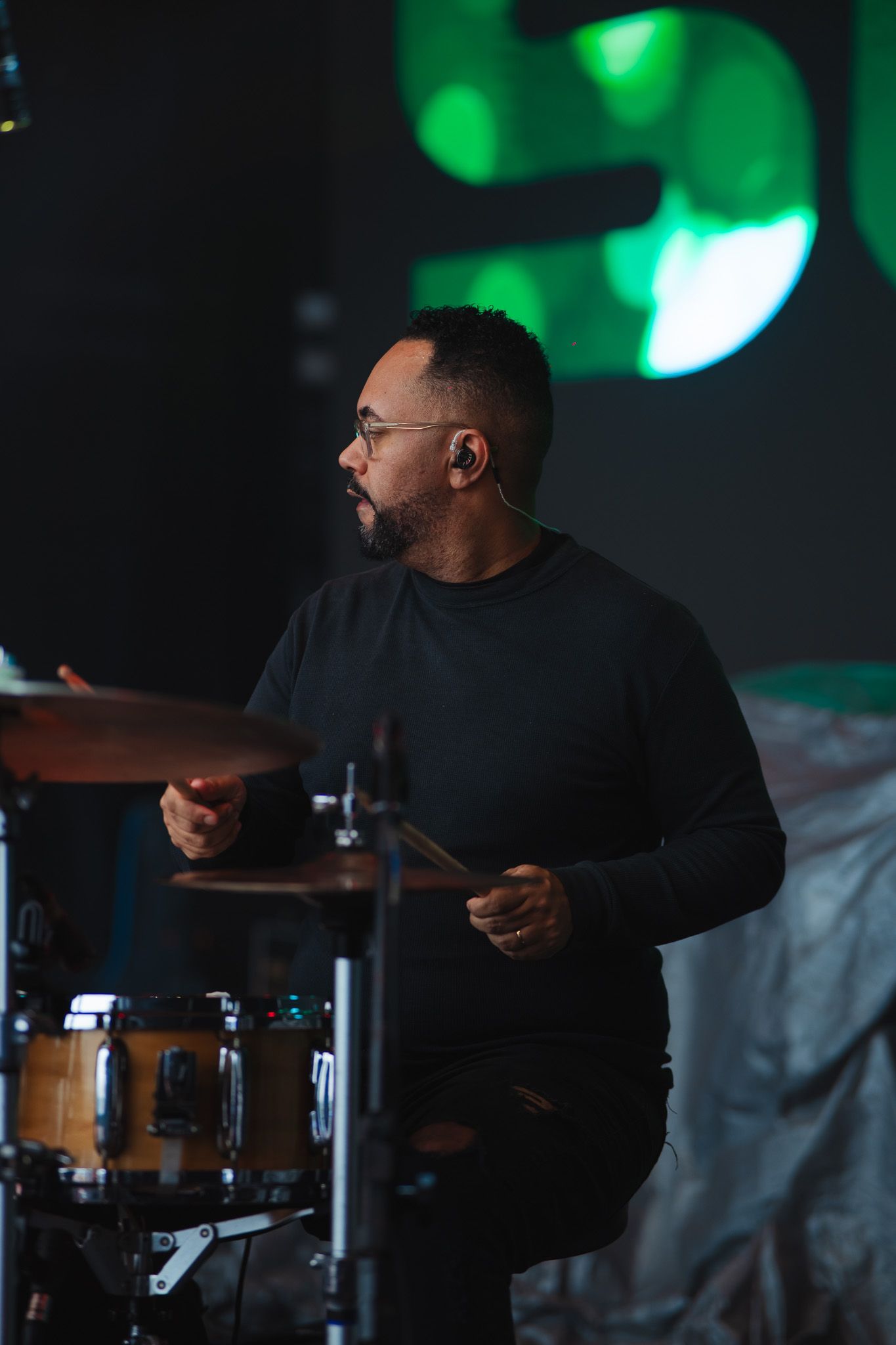 Baterista tocando bateria no palco, usando óculos e roupa preta.