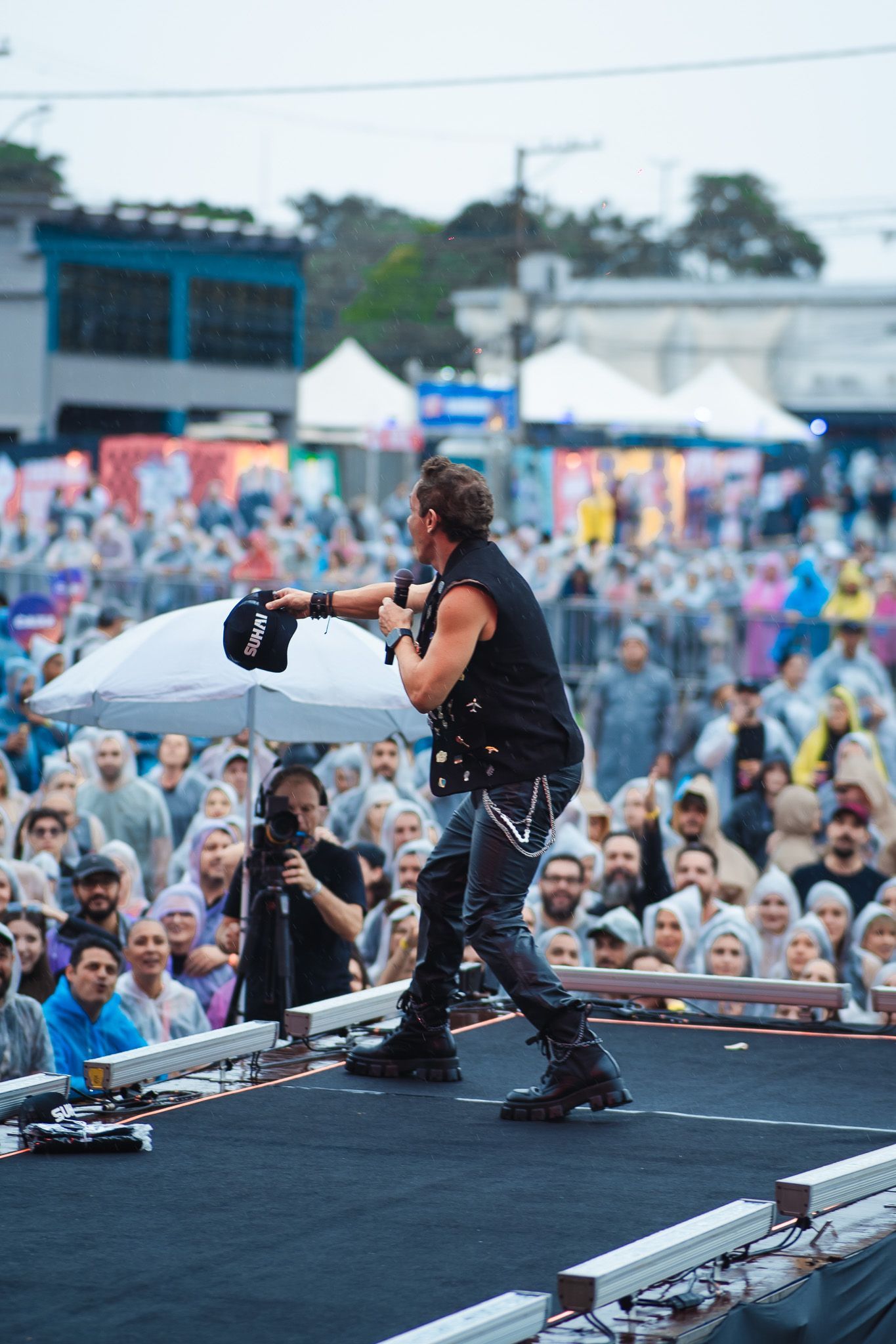 Um artista no palco com microfone, apresentando-se para uma plateia em um ambiente externo.