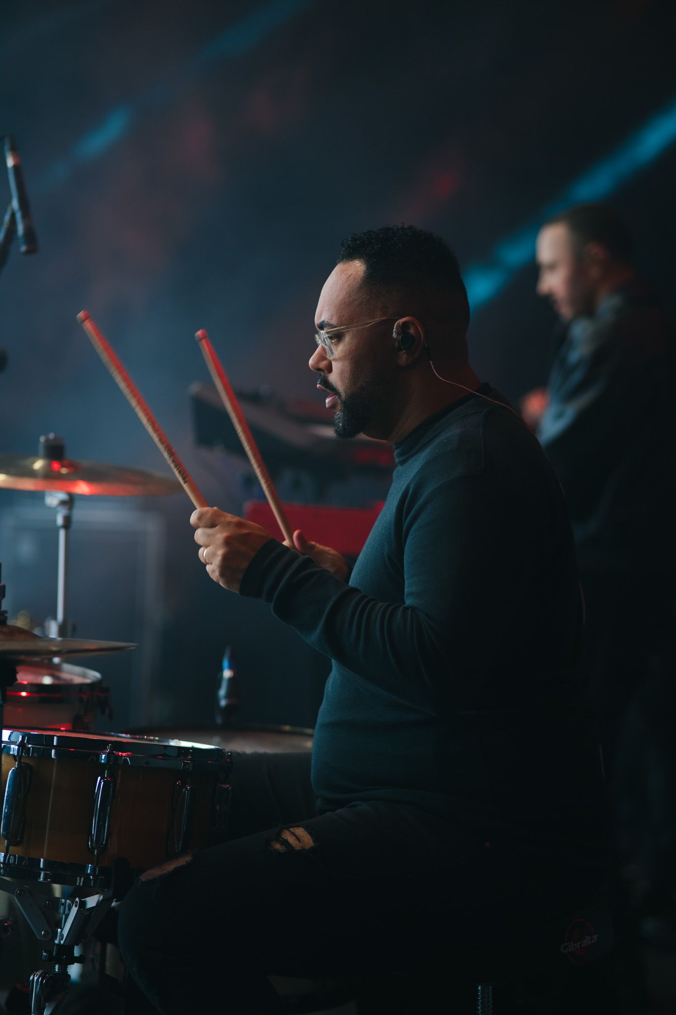 Baterista tocando bateria no palco, iluminado com luzes vermelhas e azuis. Outra pessoa ao fundo.
